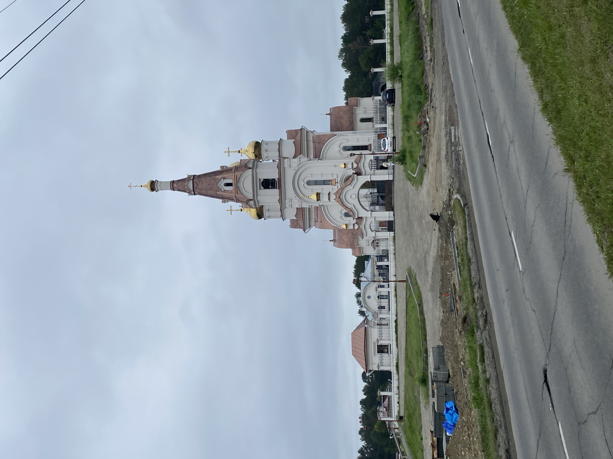 The image depicts the Church of the Kazan Icon of the Mother of God in Shelekhov, Russia, under a cloudy sky. The church is a prominent white structure with a tall, copper-colored spire topped with a golden cross. It features several smaller golden domes and arched windows. A white fence encloses the church grounds, which also include smaller white buildings with copper roofs. In front of the church, a gate with decorative ironwork is visible. Two cars, a white sedan and a dark SUV, are parked near the entrance.
The foreground of the image shows a cracked asphalt road with grass growing on its edges. Adjacent to the road, there is an area under construction with piles of dirt, concrete blocks, and a blue tarp. A few workers and some construction equipment are present in this area. The overall environment appears to be on the outskirts of a town or city, with a treeline visible in the background behind the church. The weather is overcast, suggesting a cool, possibly damp day.