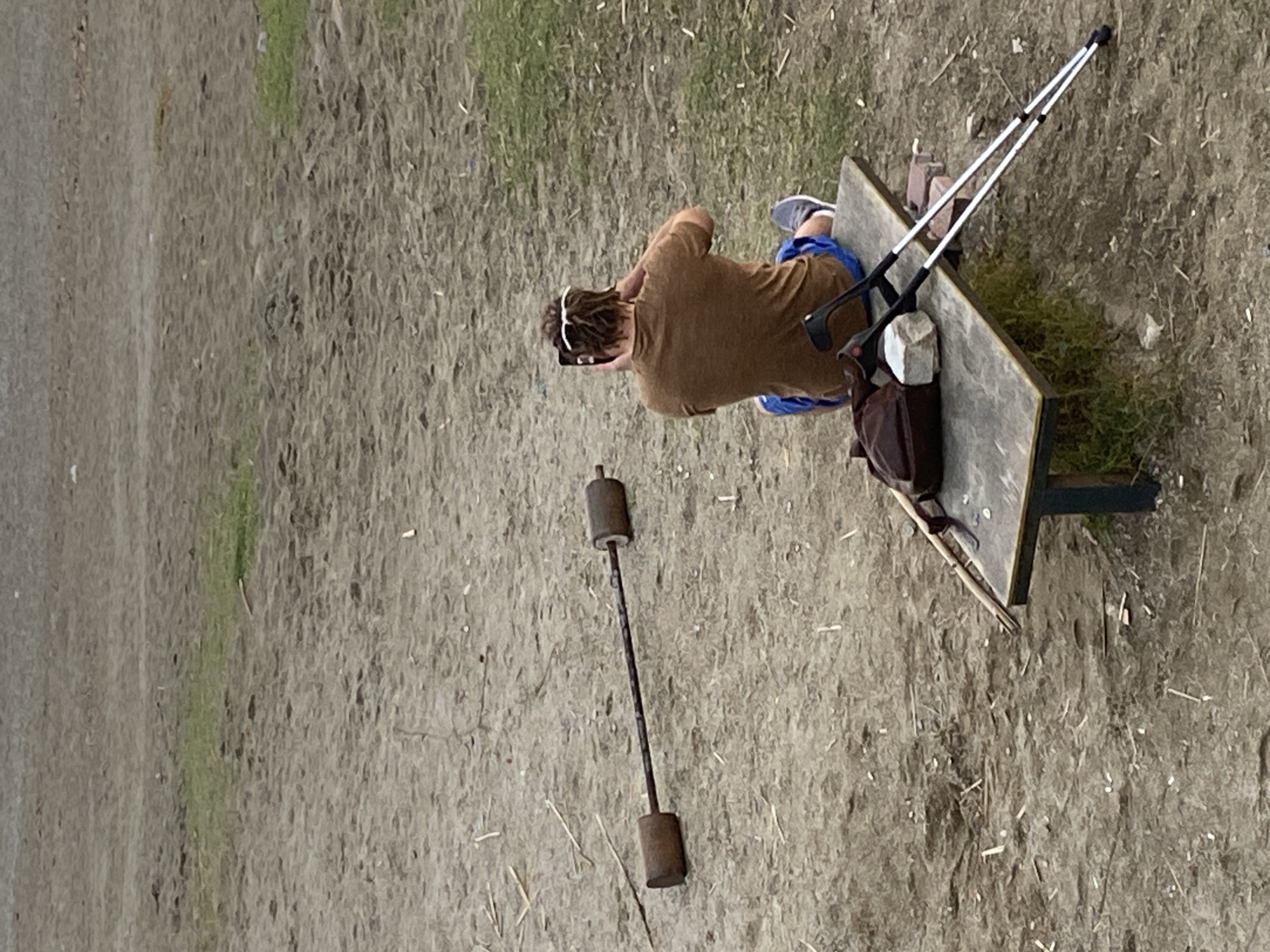 A man wearing a brown t-shirt and blue shorts is sitting on a park bench in Alanya, Turkey. He has white sunglasses perched on the back of his head and is looking at a mobile phone in his hands. The man has crutches leaning against the bench on one side, with a bag and rocks securing them in place. A short, rusted barbell lies on the ground to the side of the bench. The ground is dirt and grass, and there is a strip of green grass at the top of the image.