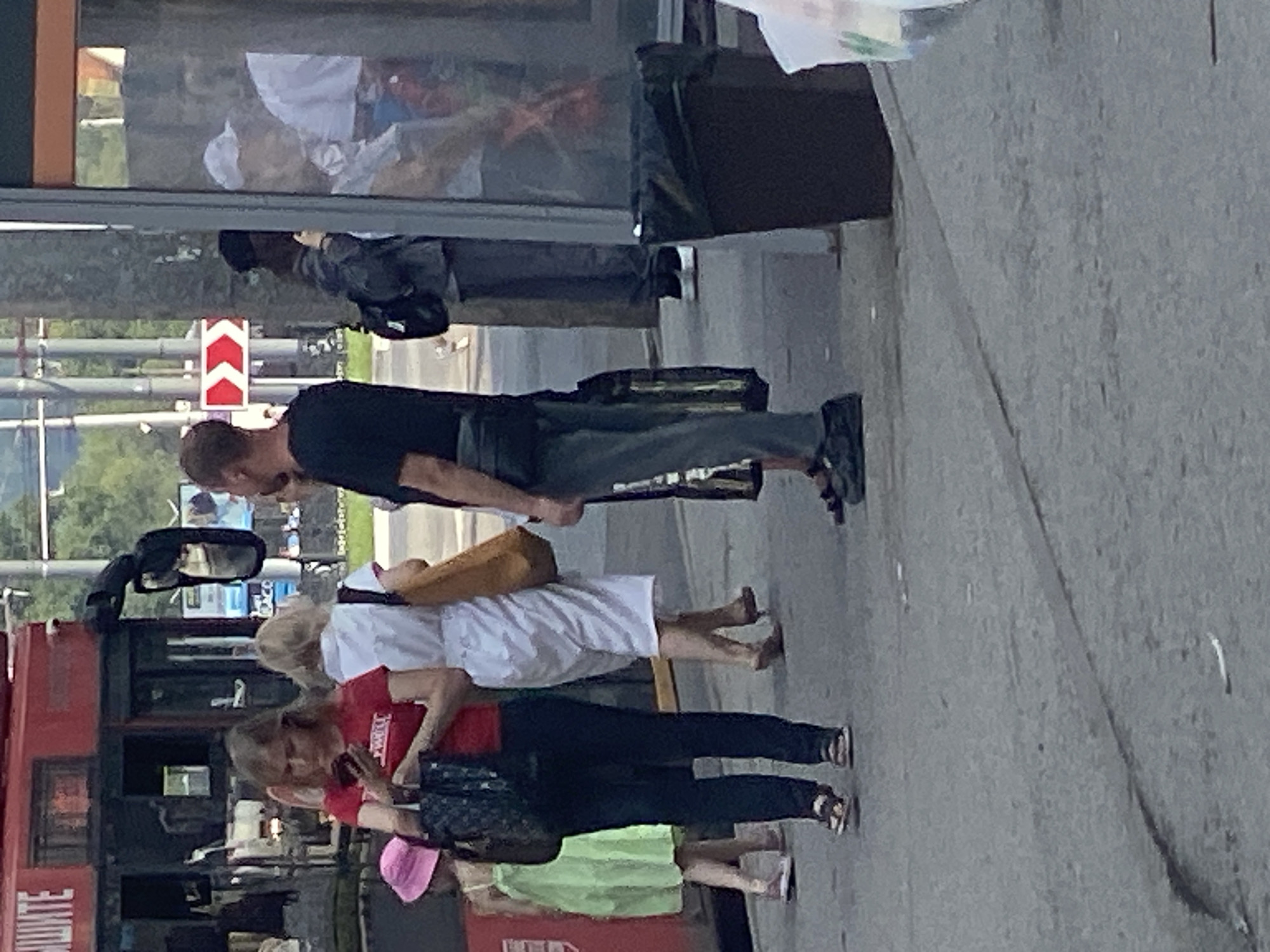 The image depicts an outdoor scene in Novokuznetsk, Russia, likely near a bus stop or public transportation hub. Several individuals are visible, some waiting, others possibly disembarking or preparing to board.

In the foreground, a woman wearing a red t-shirt and dark pants is looking at her phone. She is carrying a dark handbag. To her left, a young girl in a light green dress and a pink hat is also present. Behind the woman in the red shirt, another woman in a white dress and sandals is holding a tan bag. Further back, a man in a black t-shirt and gray pants is standing, with a black fanny pack around his waist. His stance suggests he might be waiting. To his right, another individual, partially obscured, appears to be wearing a dark jacket and backpack, and is also looking away. In the background, other figures are visible, including a person in a white cap and shirt.

The environment includes a red bus with Cyrillic lettering on the front, suggesting it's a public transport vehicle. There is also a bus stop shelter with glass panels. A black trash can stands prominently near the shelter. The ground is paved with asphalt, and there are some power poles and greenery in the background. A red and white directional sign is also visible. The lighting suggests it is daytime. The overall impression is a common urban scene involving public transportation and pedestrian activity.