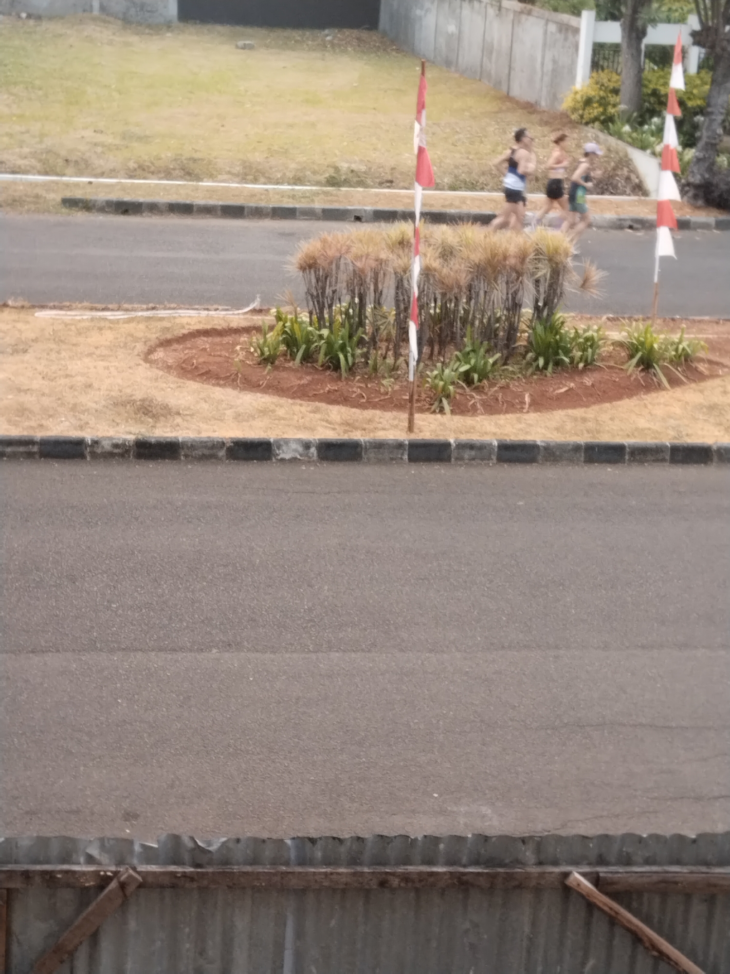 Three individuals are running on a paved road in what appears to be a residential or recreational area. Two Indonesian flags, red and white, are visible, one to the left and one to the right of the runners, planted in the ground. The area is lined with a curb and a grassy embankment, with some dry bushes and green plants. In the background, there's a concrete wall and trees, suggesting an outdoor setting. The foreground shows a paved surface and a rustic fence-like structure. The scene is likely captured from a slightly elevated position.