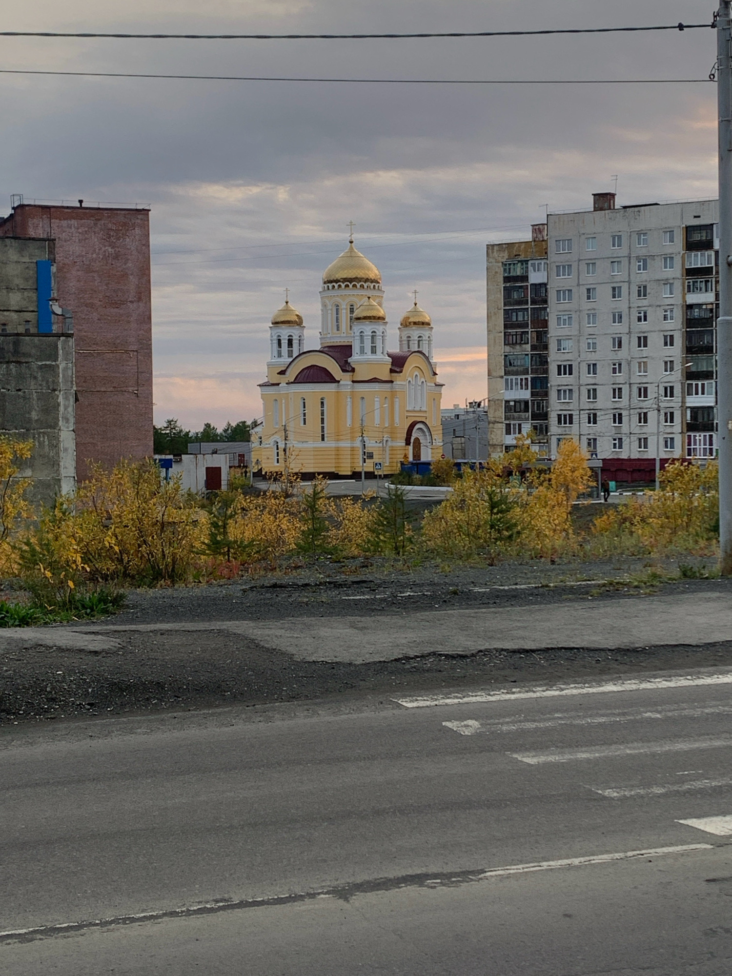 The image shows a yellow Orthodox church with golden domes, situated between two buildings in Norilsk, Russia. The church is centrally located in the frame, with a tall apartment building on the right and a brick building on the left. The foreground features a road with a pedestrian crossing. Autumn foliage in yellow and orange colors surrounds the base of the church and extends towards the foreground. The sky is overcast with clouds, and there are power lines visible across the top of the image. The overall scene depicts an urban landscape in Norilsk during the autumn season.