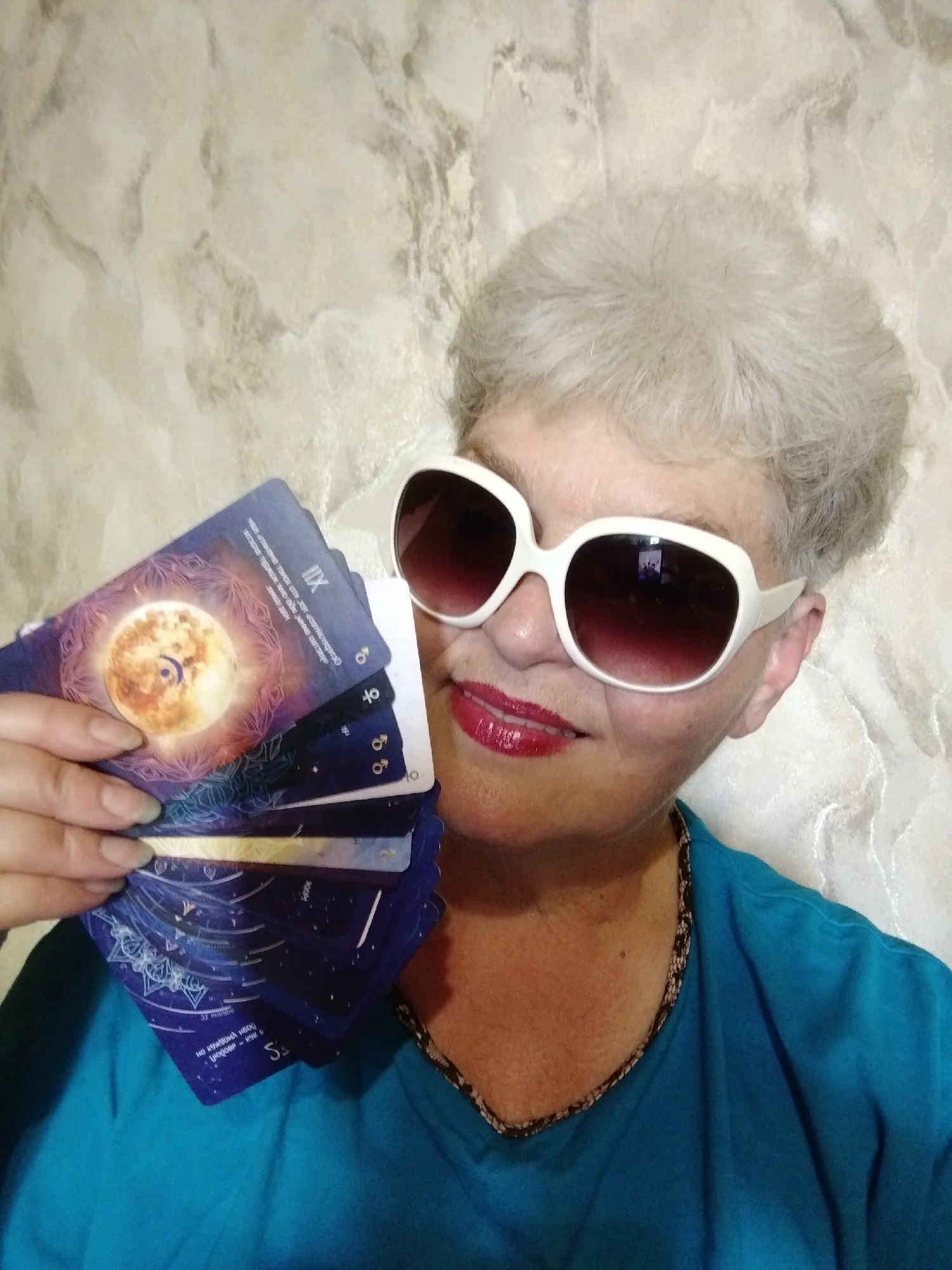 A close-up portrait features an individual, likely an older woman, with short, curly gray hair, looking directly at the camera with a slight smile. She wears large white-framed sunglasses with dark lenses, reflecting a small, indistinct image. Her lips are adorned with bright red lipstick. She is dressed in a teal or turquoise V-neck top with a dark, patterned trim along the neckline.
In her left hand, held up and fanned towards the viewer, she presents several dark blue cards, which appear to be oracle or astrological cards. The most prominent card features a golden, glowing full moon or celestial body with a crescent moon symbol inside, surrounded by intricate patterns and text. Another card clearly displays the Roman numeral "XII." Other visible cards show various astrological or planetary symbols, including "♂" (Mars) and "♀" (Venus), along with partially obscured text. Visible text from the cards includes "MODE," "S," and phrases in Cyrillic such as "боди умоджен н," "тенок оти затв," and "KEK NHO DÒN ние овтовур."
The background is a light-colored wall with a mottled or marble-like pattern. The activity suggests a presentation or reading of the cards. The image was captured indoors in Horishni Plavni, Ukraine.
