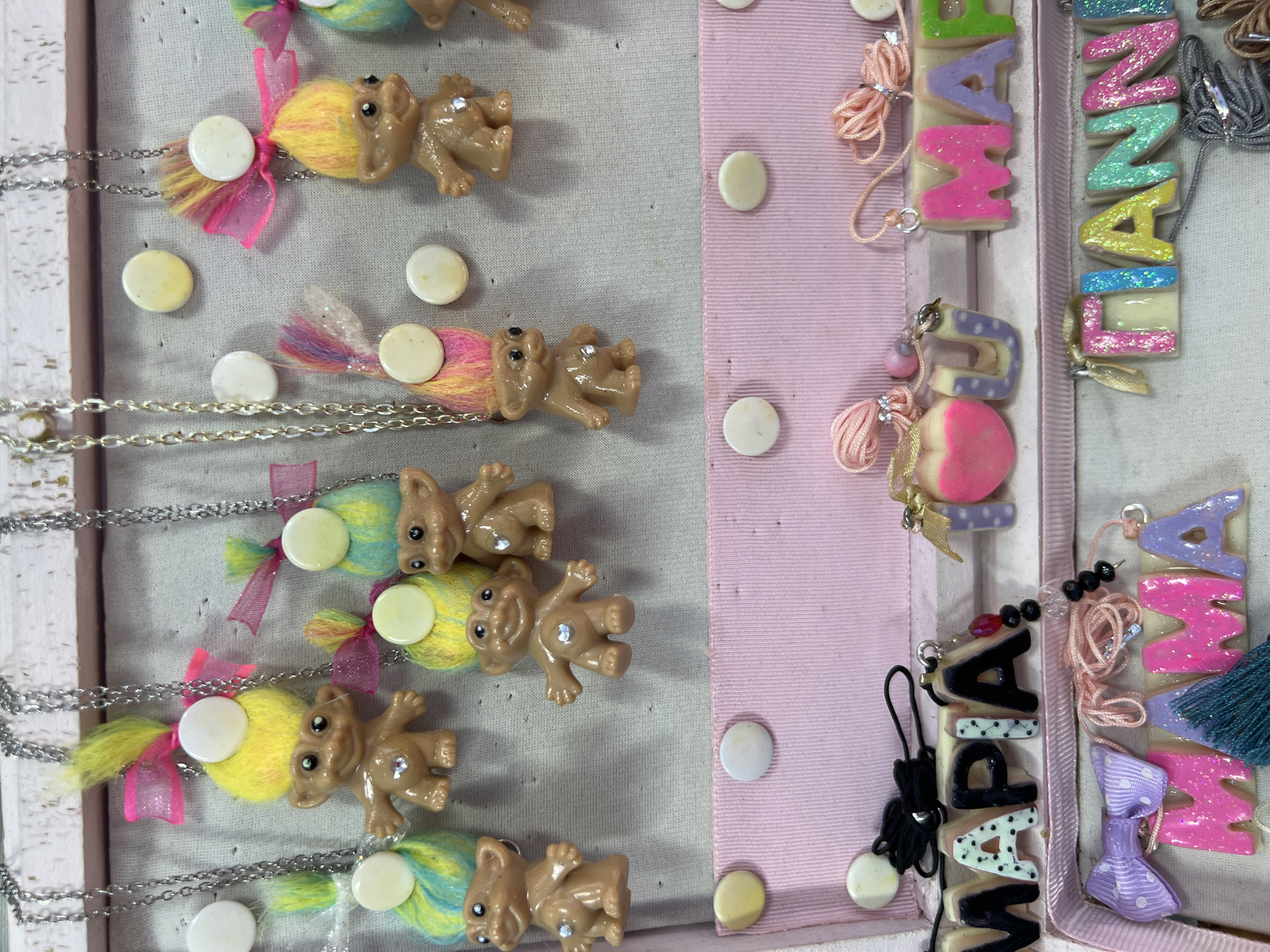 The image displays a close-up, eye-level view of a display of handmade jewelry, likely in a market or craft fair setting. The jewelry consists of necklaces featuring troll dolls with colorful hair and small gem accents on their chests, as well as letter pendants spelling out names. The display is organized on a pink tiered stand with white circular embellishments. The upper tier showcases troll doll necklaces, each suspended from a silver chain. The lower tiers display the letter pendants, some with glitter and vibrant colors, spelling "MARIA," "MAMA," and "GIANNI." The background is a textured white surface, possibly a wall or fabric. The overall impression is one of a colorful and whimsical collection of personalized jewelry. There are no people visible in the image, and the setting is not specifically identifiable as Eretria Municipal Unit, Greece.