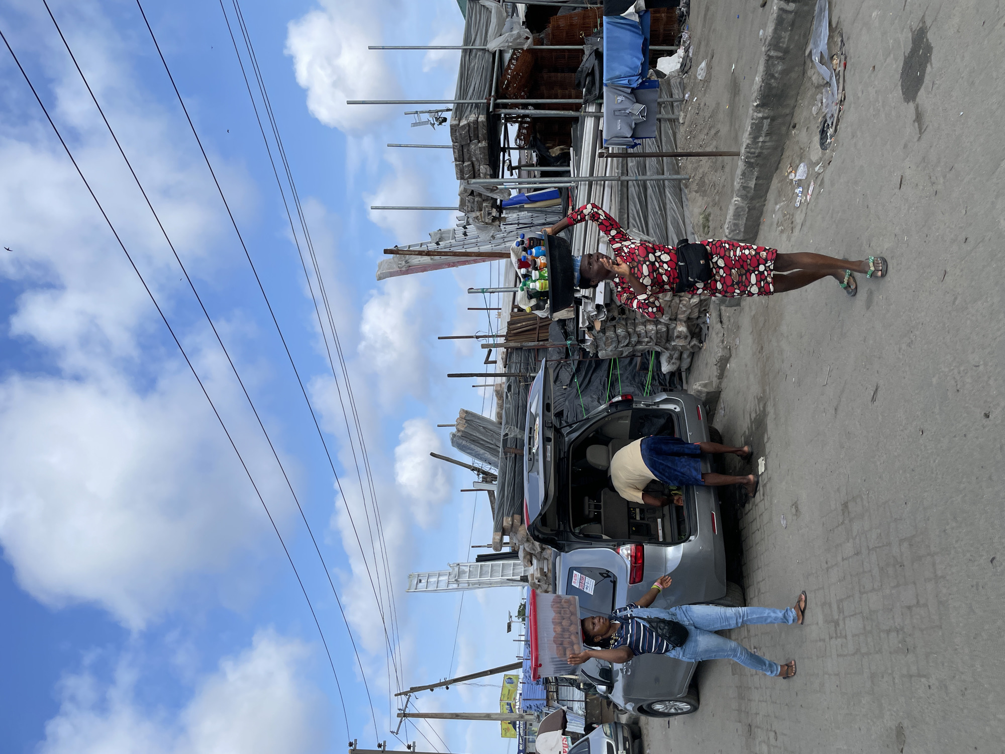 In Lekki, Nigeria, on a paved outdoor market street under a cloudy sky, two women are captured in motion. The woman on the right, wearing a red and white patterned dress and green flip-flops, balances a circular tray of colorful bottled drinks on her head. She carries a black fanny pack around her waist. To her left, a second woman in blue overalls, a striped shirt, and brown flip-flops, walks away from a gray minivan. She has a dark shoulder bag. Inside the open rear of the minivan, a man in a tan t-shirt and blue shorts is bent over, appearing to be retrieving or storing items. The background is filled with stacks of building materials, including pipes, metal bars, and bundled planks, suggesting a construction or hardware supply area. Overhead, power lines stretch across the frame.