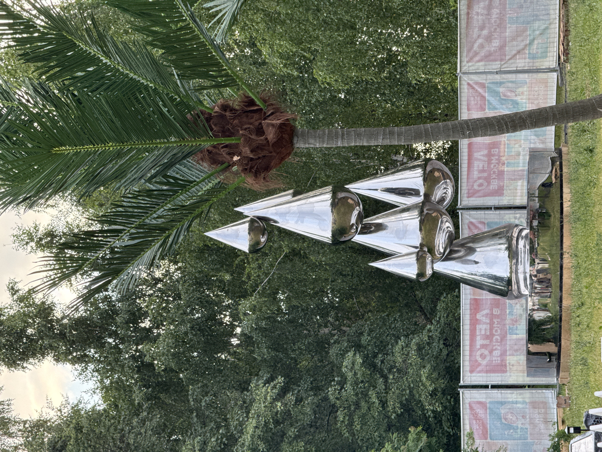The image shows a silver, cone-shaped sculpture in a park-like setting. The sculpture consists of several interconnected conical shapes, with the topmost cone featuring a rounded tip. The surface of the sculpture is highly reflective, mirroring the surrounding trees and sky. To the right of the sculpture, a palm tree with a fibrous trunk and long, green fronds is prominently featured. In the background, there are dense green trees and a white construction fence with red and blue panels displaying the word "VELO" and Cyrillic text. The ground in the foreground is covered with green grass and some scattered debris. The sky above is overcast. This sculpture is located in the area of Gorky Park in Moscow, Russia. There are no people visible in the image.