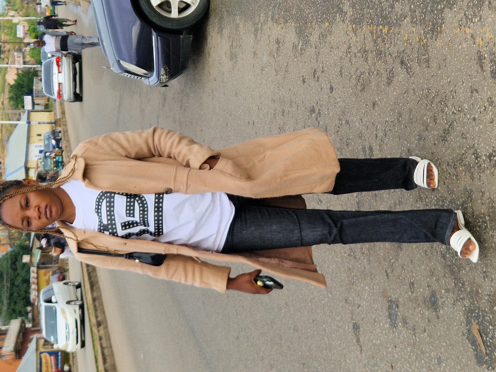 A young woman with braided hair walks on a paved street in Kabong, Nigeria. She wears a tan trench coat over a white t-shirt with a black and white graphic print, dark wash jeans, and white slide sandals. She is holding a black smartphone in her right hand. A black purse with a strap is slung over her left shoulder. In the background, several cars are visible, including a white minivan, a silver sedan, and a dark blue sedan. The environment appears to be a commercial or residential street with buildings and a hill in the distance.