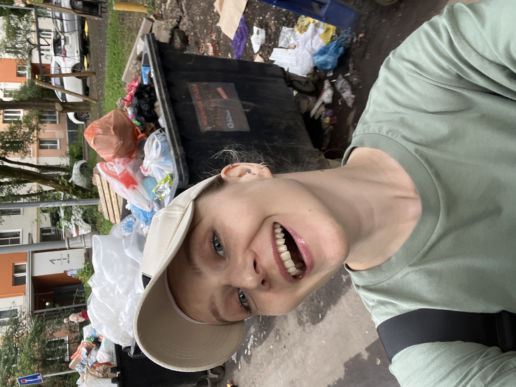 A young woman with blonde hair and blue eyes, wearing a light-colored baseball cap and a light green t-shirt, smiles broadly at the camera. She has a pierced ear with two hoop earrings and a silver stud. Behind her, several overflowing garbage dumpsters are visible, with trash scattered around them. In the background, a residential building with orange and white facades, trees, and parked cars are present. The scene appears to be a residential courtyard in the city of Khimki, Russia.
