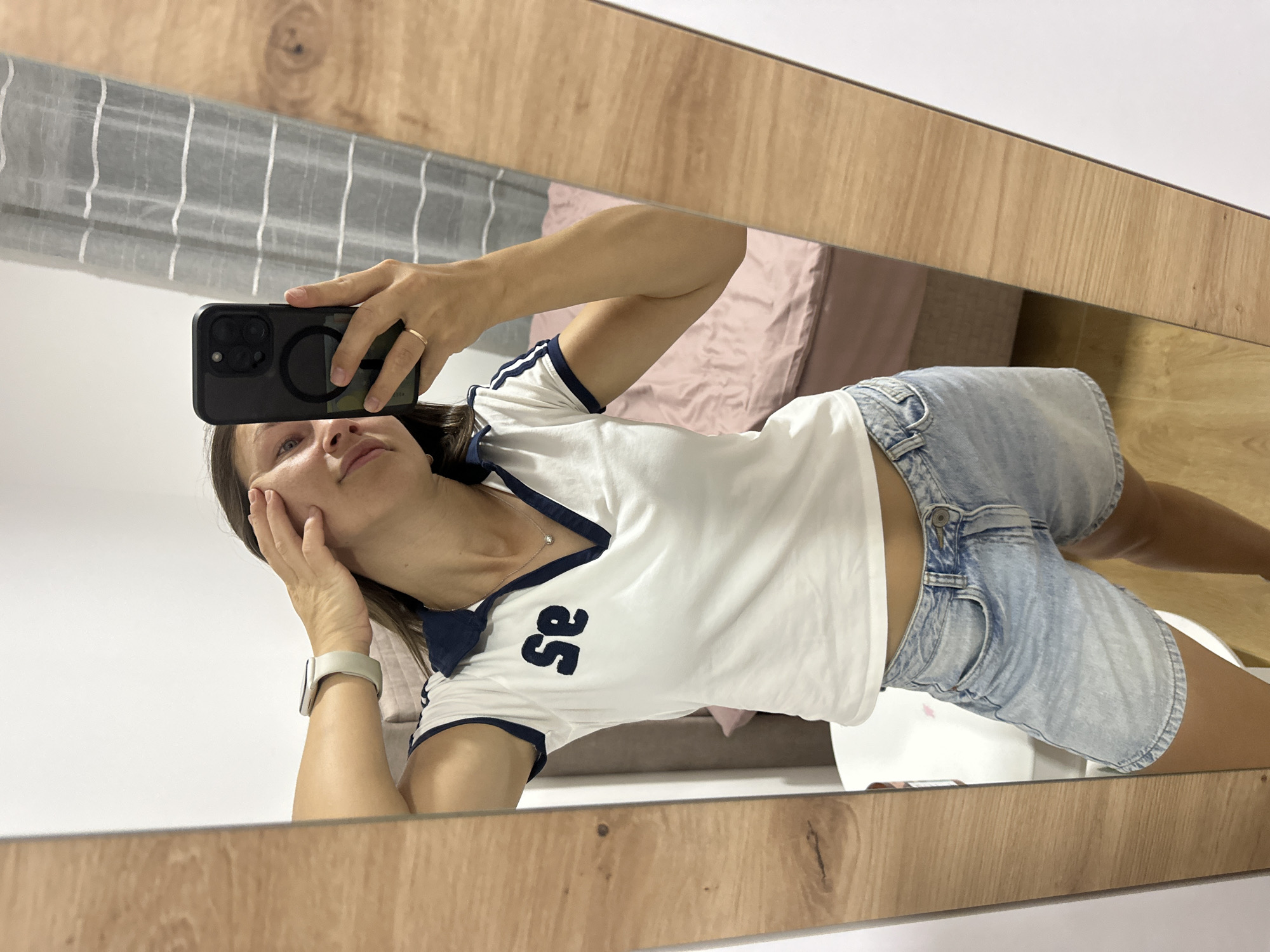 In an indoor setting, possibly a bedroom in Donji Pobori, Montenegro, a woman is taking a mirror selfie. She is wearing a white polo shirt with navy blue trim and the letters "se" in navy blue on the chest, paired with light-wash denim shorts. She has a smartwatch on her left wrist and a ring on her right ring finger. The woman is leaning forward slightly, with her right hand touching her face near her eye, and her left hand holding a black smartphone, positioned to capture the reflection. The background includes a bed with a pink duvet, a white curtain with gray stripes, and a wooden-framed mirror. The floor appears to be light-colored wood.