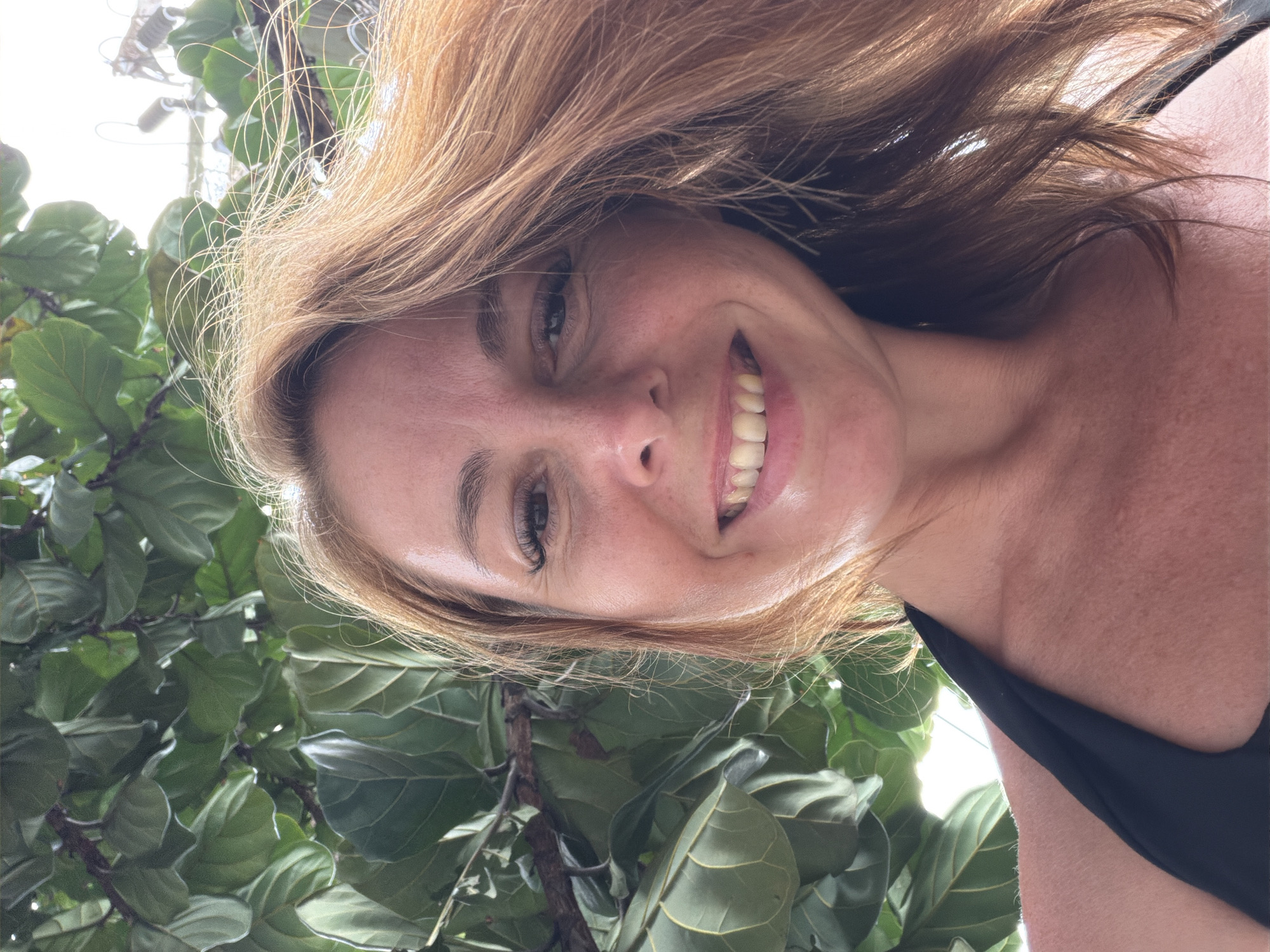 A woman with auburn hair and fair skin smiles at the camera from a low-angle perspective. She is wearing a black bikini top. Large, green leaves of a Fiddle Leaf Fig tree are visible in the background, along with a hint of a bright sky and some man-made structures, suggesting an outdoor setting, possibly a garden or resort in a tropical location like Ban Bang Rak, Thailand.