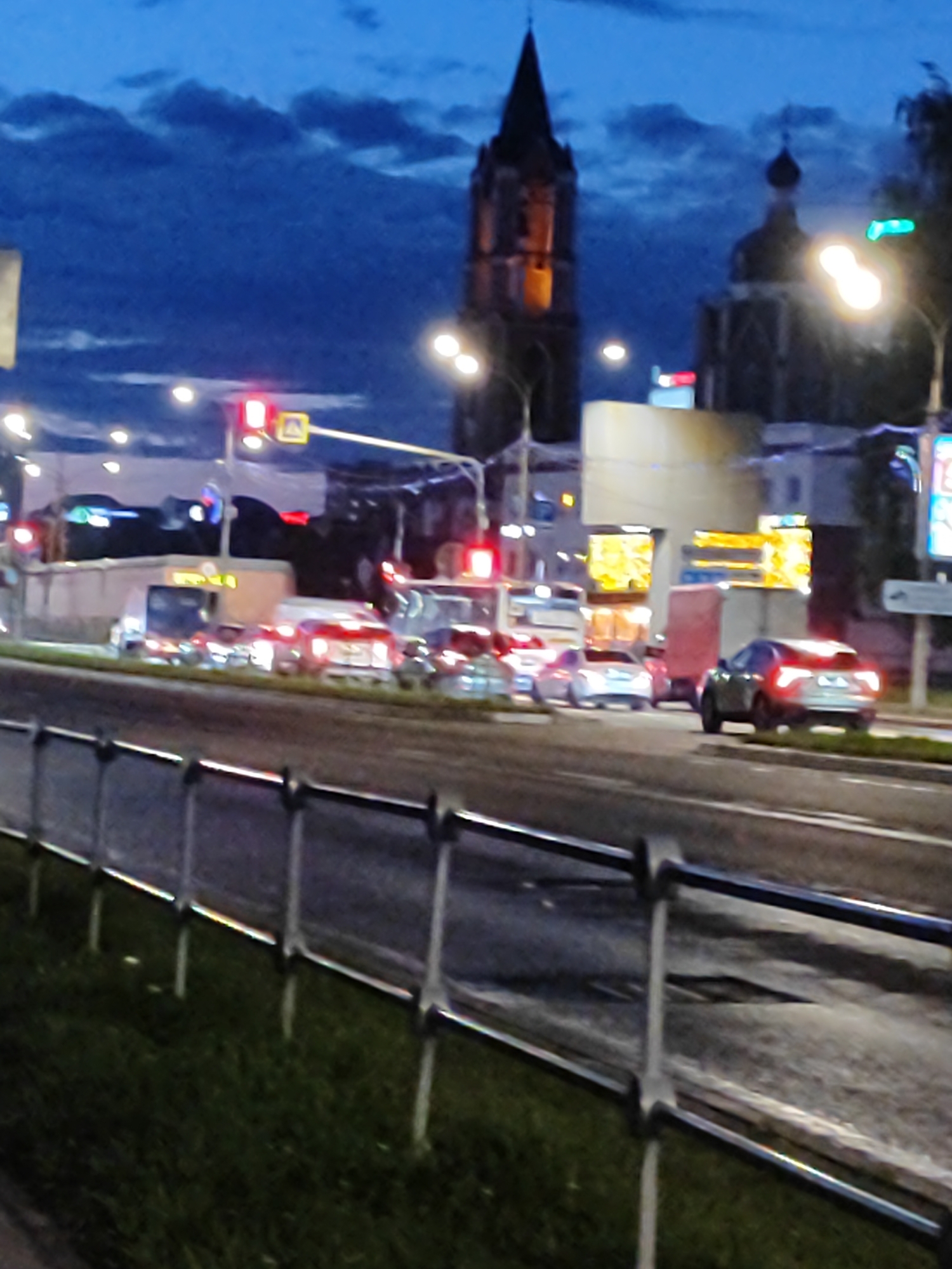 The image captures a street scene at dusk in an urban environment. A traffic light displays a red signal, indicating that vehicles should stop. Several cars are stopped or moving slowly in traffic. In the background, buildings and a tall, pointed structure resembling a tower or church spire are visible. Streetlights illuminate the scene, and the sky is a deep blue with some clouds. The foreground features a metal railing and grass.