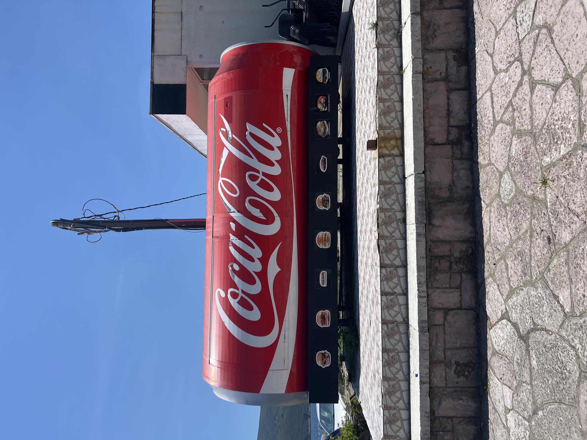 The image shows a large, cylindrical Coca-Cola can replica serving as a roadside food stand in Baošići, Montenegro. The can is predominantly red with the white Coca-Cola logo prominently displayed. Along the bottom of the can, there are several framed pictures of food items, including burgers, hot dogs, and kebabs, suggesting the types of food offered. The stand is situated on a raised platform made of stone and tiled steps. In the background, a white building with a dark square window is visible, along with stacks of chairs. Beyond that, a body of water and a lush, green hillside are present under a clear blue sky. The overall scene depicts an outdoor dining or food service establishment in a coastal area.