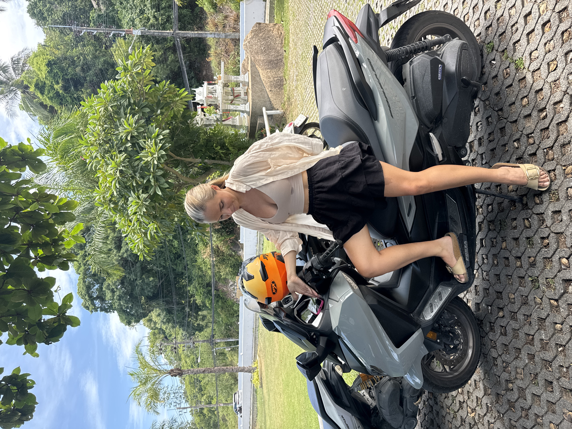 A blonde woman sits on a gray scooter in what appears to be an outdoor parking area in Ko Samui District, Thailand. She is wearing a beige crop top, a light-colored open shirt, and a black ruffled skirt. Her right leg is bent, resting on the scooter. A bright orange and yellow helmet is placed on the scooter's handlebars. The background shows lush green trees, a blue sky with scattered clouds, a small white shrine structure, and a road with a car in the distance. The ground is paved with interlocking hexagonal stones.