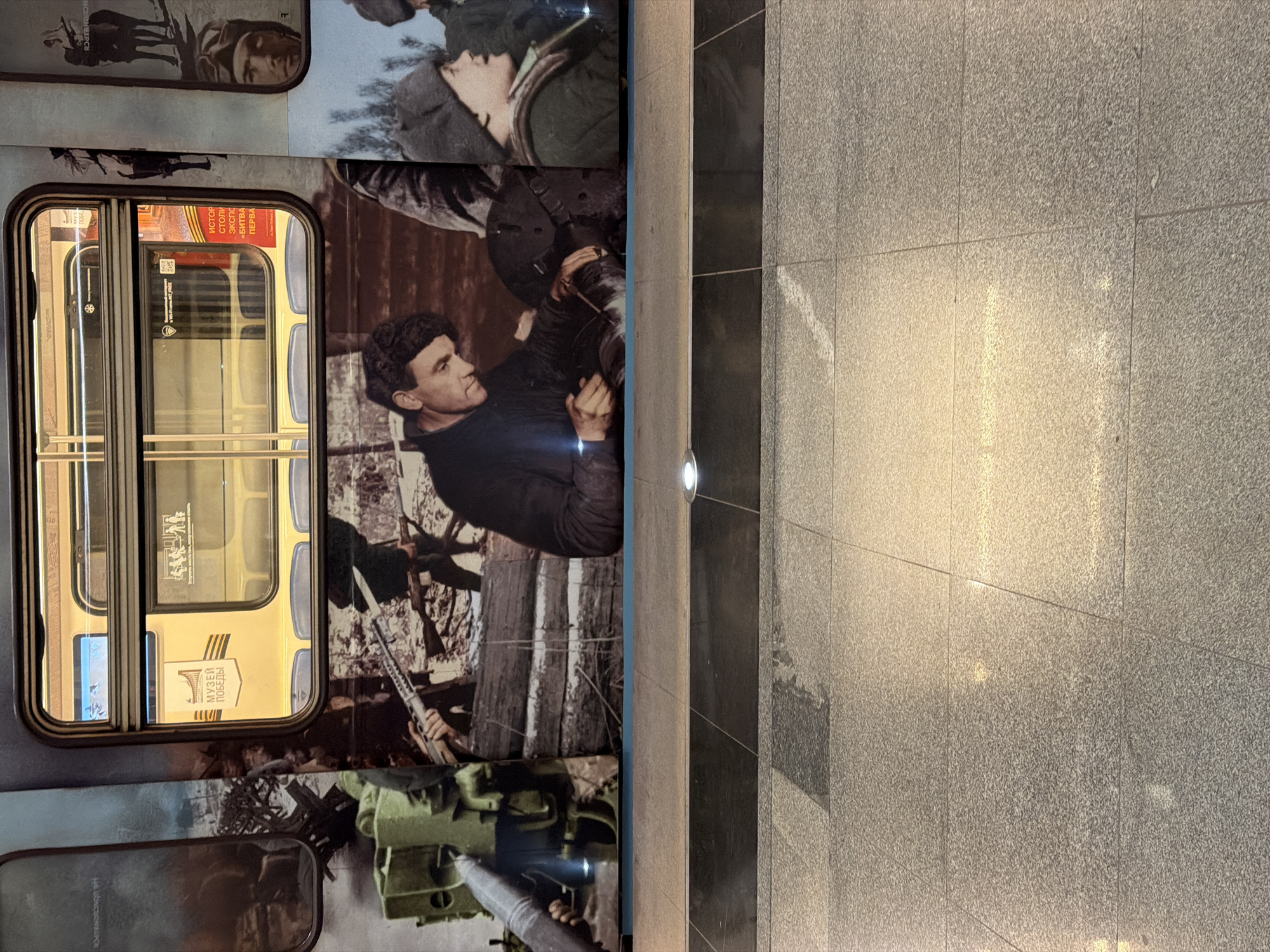 The image shows part of a wall with a mural featuring historical imagery, next to a tiled floor in what appears to be a public space in Moscow, Russia. The mural includes images related to military history, including figures in military uniforms and equipment, and the reflection of a modern subway car within a historic train. The tiling is gray and shows signs of wear or weathering.
