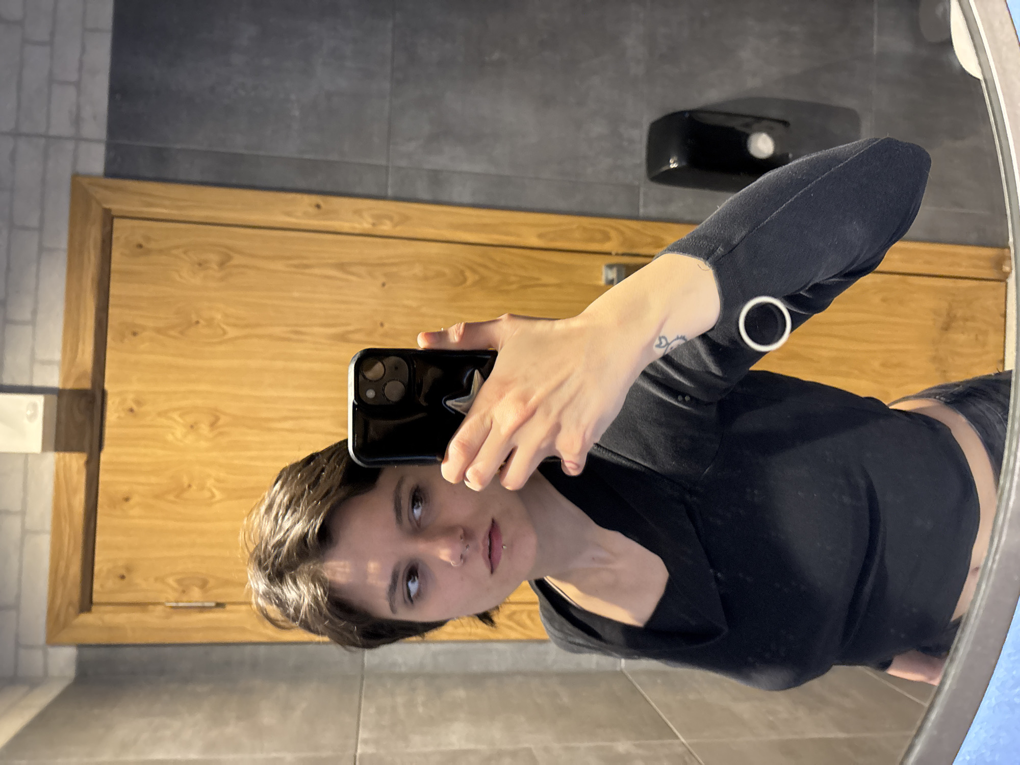 A woman is taking a selfie in a bathroom. She is holding a black smartphone in her right hand, and her face is visible in the reflection of the mirror. She has short brown hair and a nose and lip piercing. She is wearing a black top with a round button on the sleeve.
The bathroom has a light brown wooden door behind her, and the walls are tiled in gray. A black soap dispenser is mounted on the wall above the door. The floor is tiled with a different pattern of gray tiles.
Without additional information or landmarks in the image, determining the specific location within Sofia, Bulgaria is not possible.
