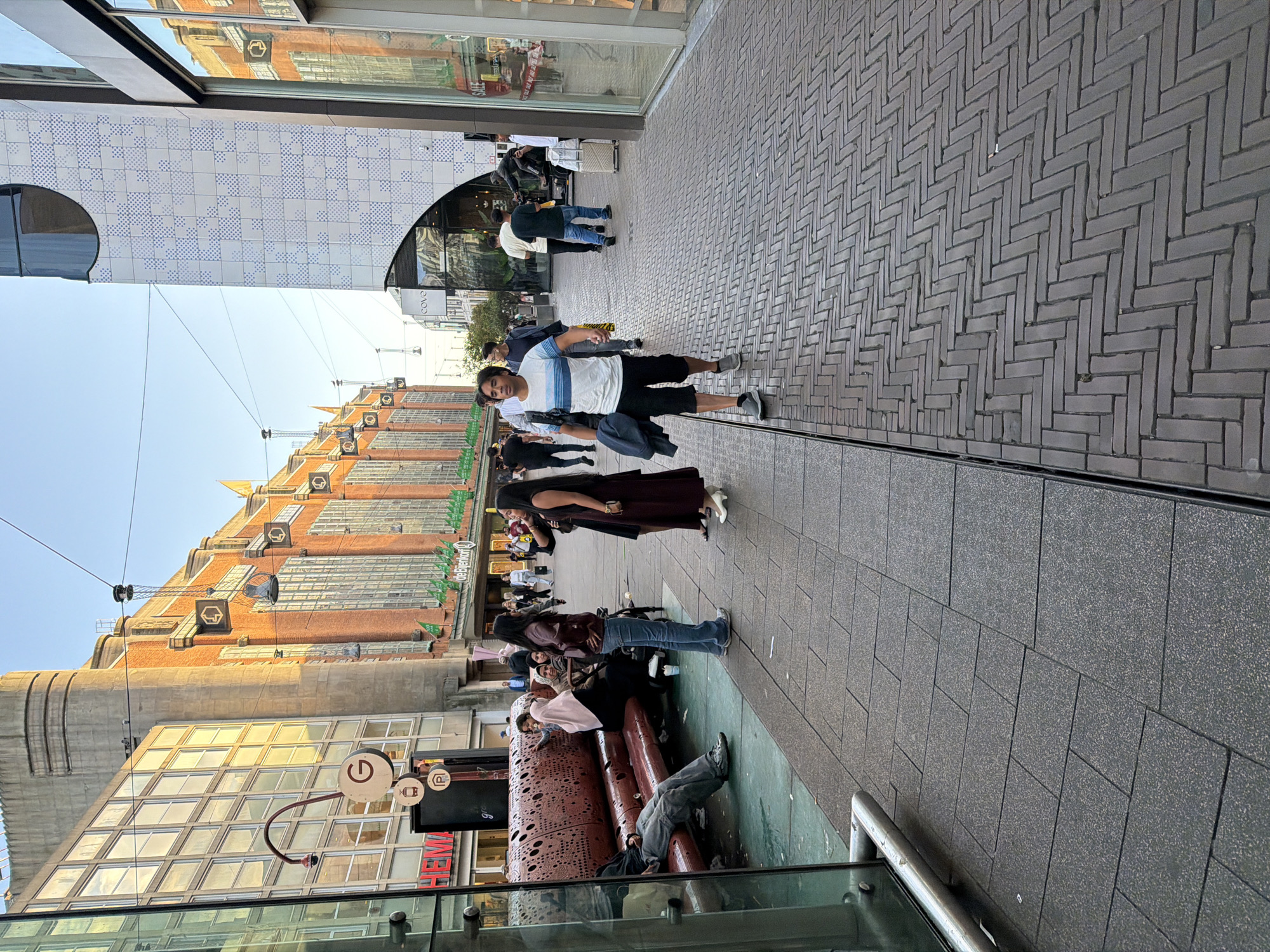 The image depicts a street scene in The Hague, Netherlands. The foreground shows a brick-patterned walkway with a reflective surface, possibly wet. Several people are visible walking and standing along the street. In the middle ground, a man in a striped shirt and shorts walks towards the camera. Further back, a woman in a dark dress is standing, and another woman in a maroon top and jeans is walking. A group of people are gathered near a circular red sculpture with intricate cutouts. In the background, the facade of a building with orange brickwork and large windows is visible, as is a building with a white tiled exterior featuring a geometric pattern. The scene appears to be outdoors during the daytime, with a clear sky. The overall atmosphere suggests a bustling urban environment. There is text visible on a sign that reads "HEMA".