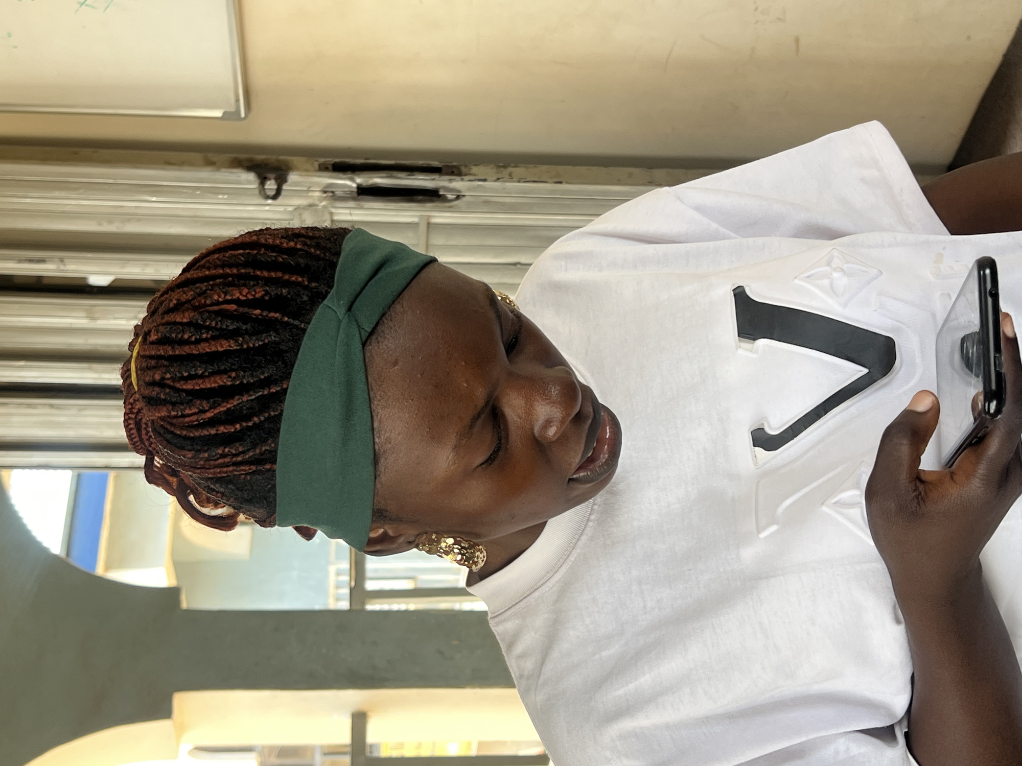 A young Black woman with dark skin, braided hair tied back with yellow bands, and a green headband is looking down at a smartphone she holds in her right hand. She is wearing a white t-shirt with a raised black and white logo of the letters "LV" and a pattern of four pointed diamonds below it. She has large gold hoop earrings. She appears to be outdoors, with a blurry background of architectural elements, possibly arches and windows, suggesting a setting in Jos, Nigeria. The lighting is bright, indicating daytime.
