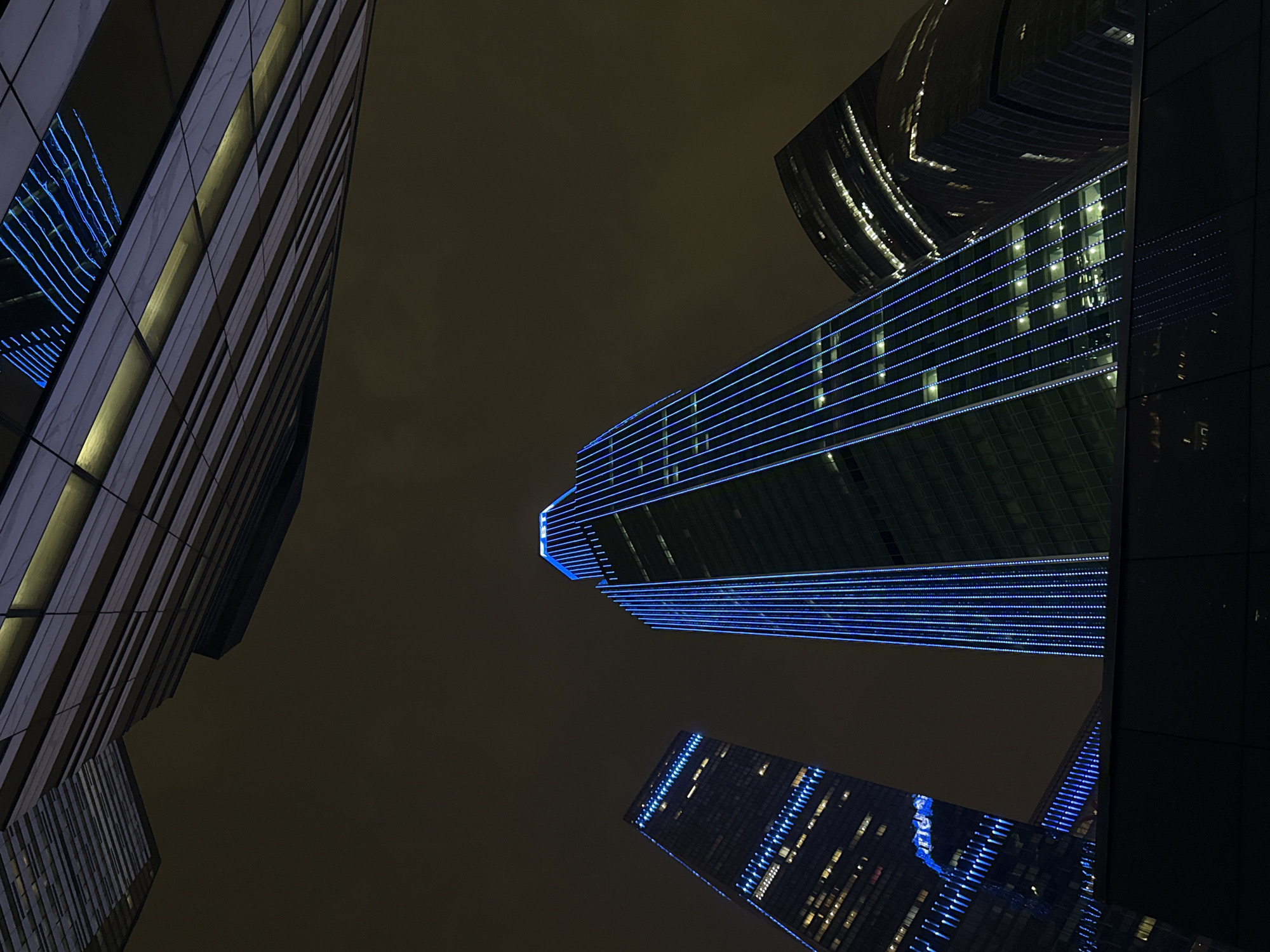 The image captures a night view of several modern skyscrapers in Khimki, Russia, seen from a low angle looking upwards. The sky is dark and overcast. The buildings are illuminated with blue lights running horizontally along their facades. One skyscraper in the center is prominently lit with these blue lines. Other skyscrapers are partially visible, with some showing reflections of the lit buildings. The architectural style is contemporary, featuring glass and steel constructions.