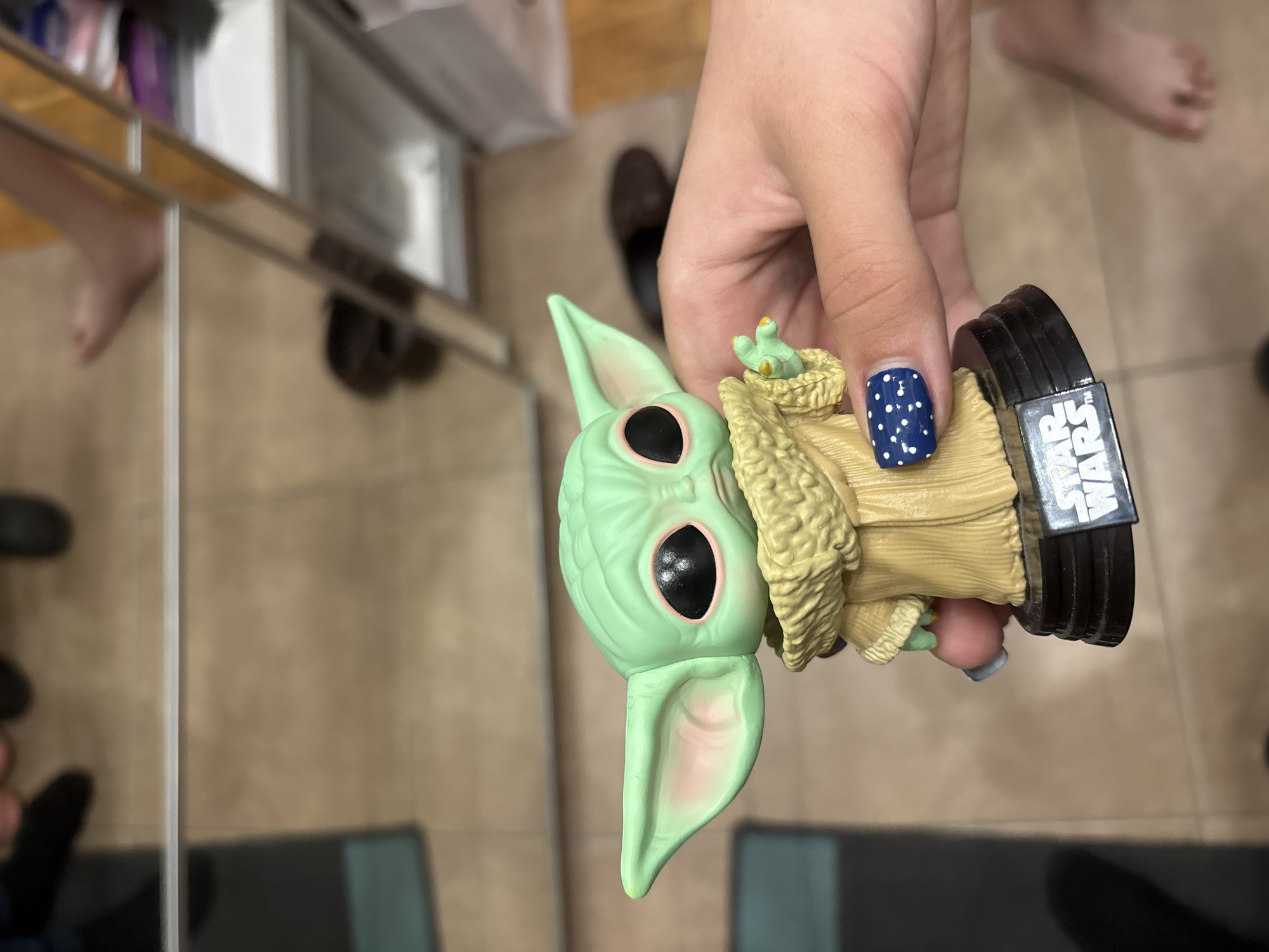 A person is holding a "The Mandalorian - Grogu" figurine against a background of a tiled floor and mirrored furniture. The person is holding the figurine by its base, which has "STAR WARS" printed on it. The figurine is green with large black eyes and wearing a tan robe. The person has dark blue nails with white polka dots on one finger. The location appears to be indoors, potentially a bathroom or dressing area. There is no visual information to suggest the location is in the city of Saint Petersburg, Russia.