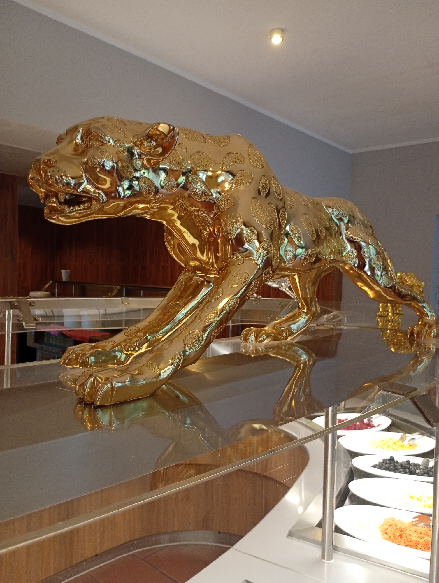 The image shows a golden sculpture of a leopard in a stalking pose. The sculpture is highly polished and reflects its surroundings. It is placed on a shiny surface, likely a buffet table, in what appears to be a restaurant or hotel setting. Behind the sculpture, there is a wooden cabinet and a glimpse of a buffet line with various food dishes in white plates. The ceiling is white with a single spotlight. This scene is located in the city of Nordhausen, Germany, likely within a dining establishment. There are no people visible in the image.