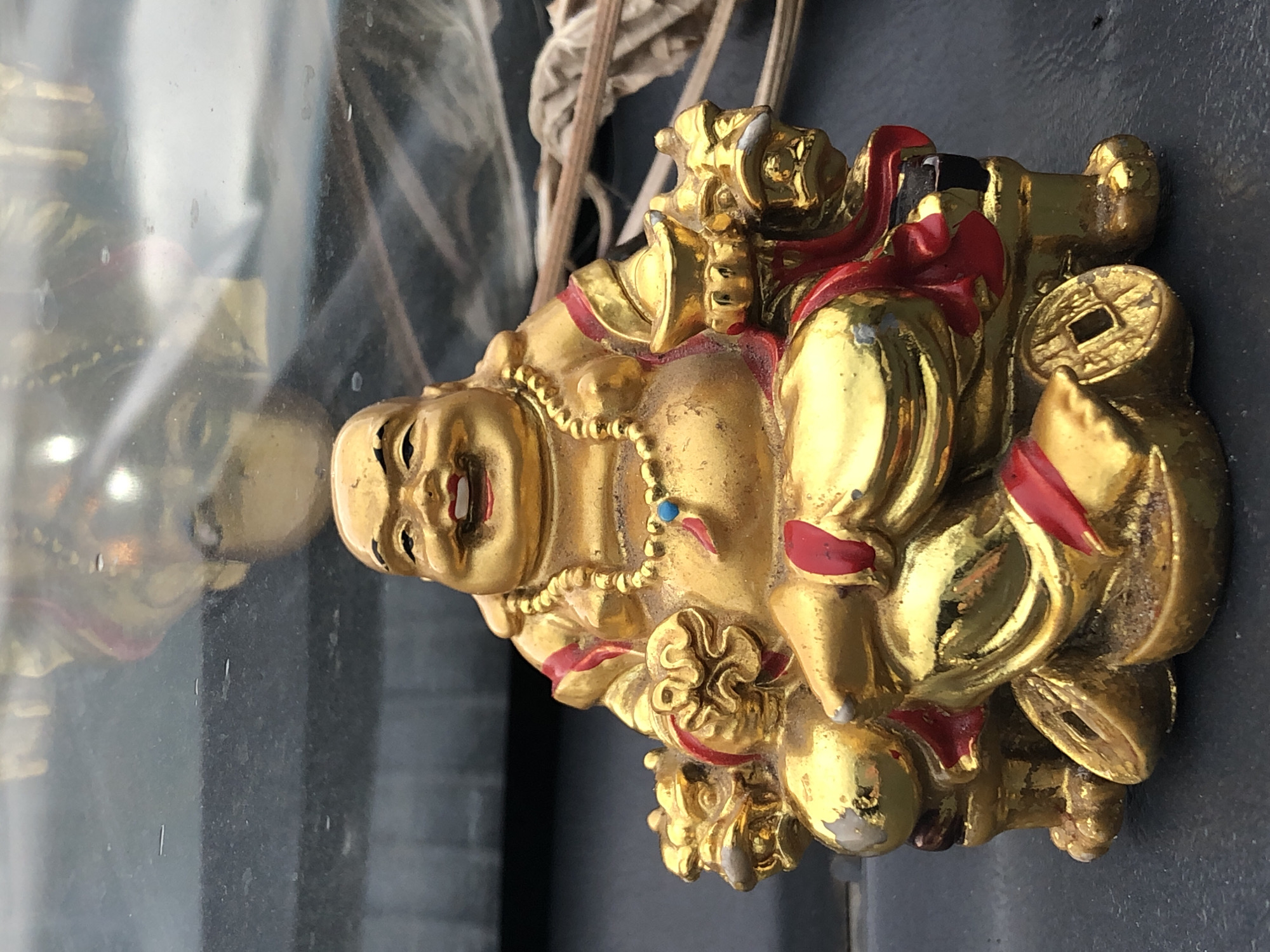 This is a close-up, slightly angled, horizontal shot of a golden, decorative figurine, likely a laughing Buddha or a similar deity. The figurine is predominantly gold-colored with accents of red and a small blue bead on a necklace. It appears to be sitting and is adorned with intricate details, including what looks like a coin with a square hole. The figurine is placed on a dark, reflective surface, possibly glass or polished stone, which shows a distorted reflection of the figurine and its surroundings. In the background, out of focus, are some dry, straw-like elements and a dark, textured surface. The lighting suggests it could be daytime, but the specific time of day, weather, or explicit location cues are not evident beyond the suggestion of an indoor setting with reflective surfaces. There is no visible text. The figurine itself seems to convey a sense of joviality and prosperity.