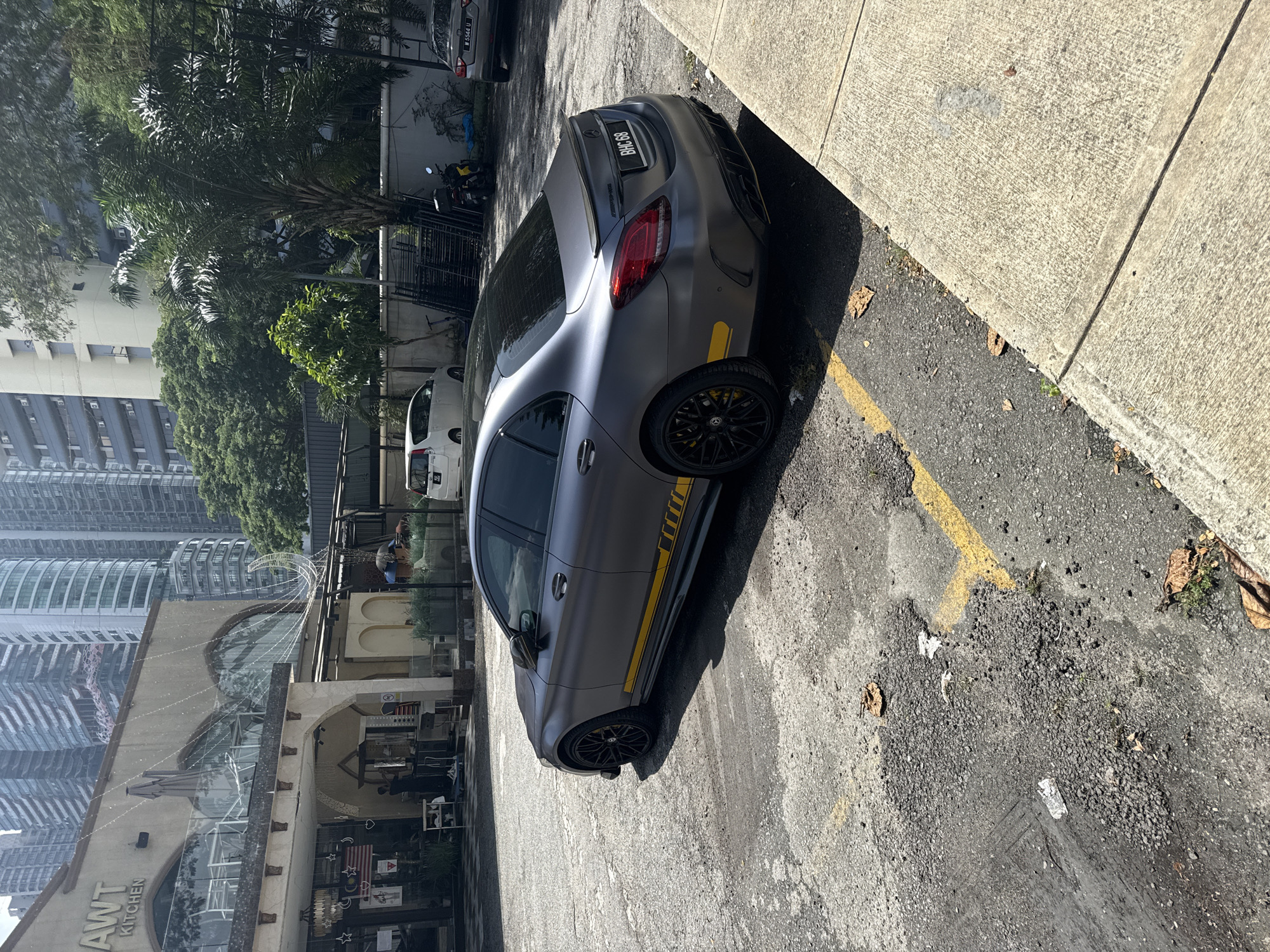 The image displays a sunny daytime street scene in Kuala Lumpur, Malaysia, centered around a dark grey, possibly matte finish, Mercedes-Benz sedan. The car is parked on an asphalt lot, seen from its rear three-quarter angle, revealing black multi-spoke wheels with yellow brake calipers and a distinctive yellow stripe decal along its lower side. The license plate "BHC 89" is visible on the rear bumper, which also features a subtle spoiler.
The parking area itself is a bit worn, with yellow markings on the asphalt, and a clean concrete sidewalk runs alongside it. In the background, a vibrant urban environment unfolds, characterized by numerous modern high-rise buildings and abundant tropical greenery, including palm trees and other dense foliage, suggesting a warm climate and metropolitan setting.
Adjacent to the parking area, a building with arched architectural elements and large windows is partially visible, bearing the sign "AWT KITCHEN" on its facade. White string lights adorn this building, and a Malaysian flag can be seen hanging near an entrance, reinforcing the location in Kuala Lumpur. Other vehicles, including a white van and a silver sedan, are partially visible in the background, along with a yellow scooter behind a fence. The scene depicts a calm, typical urban moment under bright sunlight, casting sharp shadows.