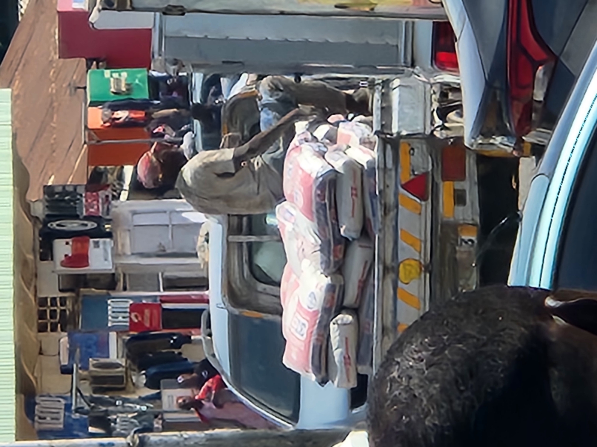 The image shows a heavily laden truck in an urban environment, likely in the city of Unknown, Unknown. The truck is carrying what appears to be a large amount of construction material, including bags that seem to contain cement, stacked high on the back.
A person can be seen amidst the load, possibly securing the cargo or preparing for unloading. Other items and objects are visible within the truck's storage area, including boxes and miscellaneous supplies.
The environment around the truck appears to be congested, possibly indicating a busy marketplace or construction site. A portion of a car or vehicle is present in the foreground, partially obscuring the view of the truck. The scene is characterized by a sense of activity and potential urban development.