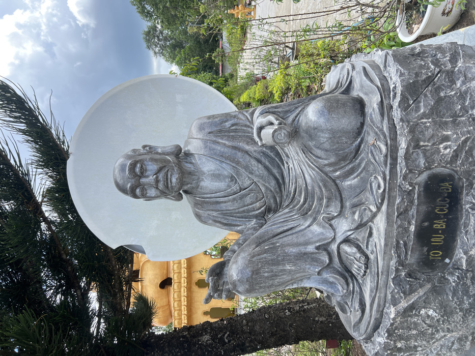 A dark grey stone statue of an elderly, bald male figure, identified by the inscription "TO UU-BA-CUC-DA" on its pedestal, is prominently displayed outdoors in the city of Đăk Mar, Vietnam. The figure is seated cross-legged, draped in robes, with a stern, wrinkled face, a beard, and elongated earlobes. His right hand extends forward with an open palm, while his left hand holds a rolled scroll on his knee. A large, smooth, light grey circular disk serves as a backdrop behind his head. To the left, a large coniferous tree partially obscures a golden-orange, multi-tiered temple structure in the background. To the right, a cultivated garden area with various green plants and small trees extends along a light-colored path. The sky above is overcast with grey clouds.