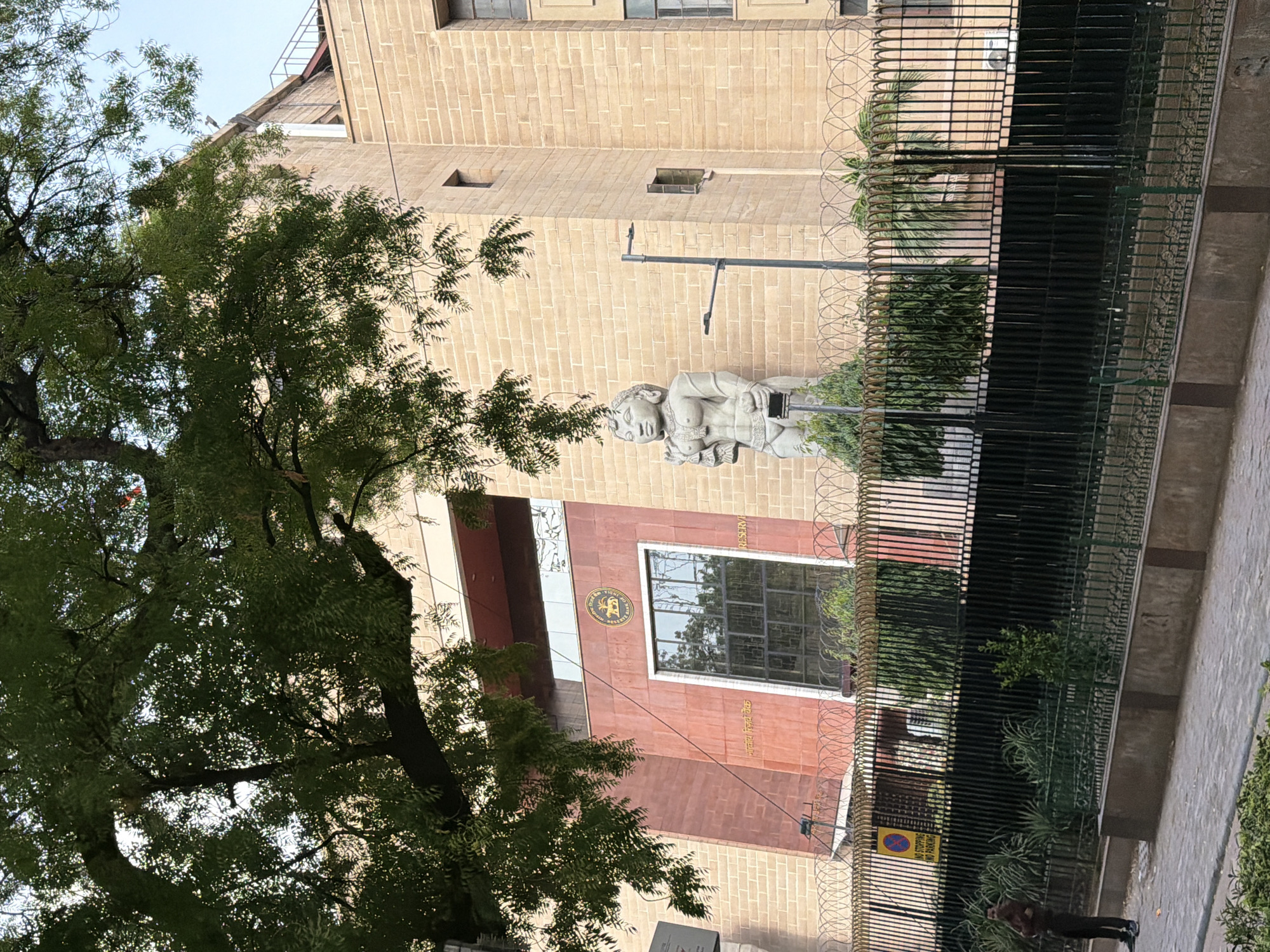 The image shows the Reserve Bank of India building in New Delhi, India. The building's facade is made of beige brick with some reddish-brown sections. A large statue of a female figure stands outside the building, to the left of a large window. A fence with barbed wire on top surrounds the property. A tree with green leaves partially obscures the top of the building. A person in a dark shirt and pants stands near a "No Stopping No Parking" sign on the left side of the frame.
