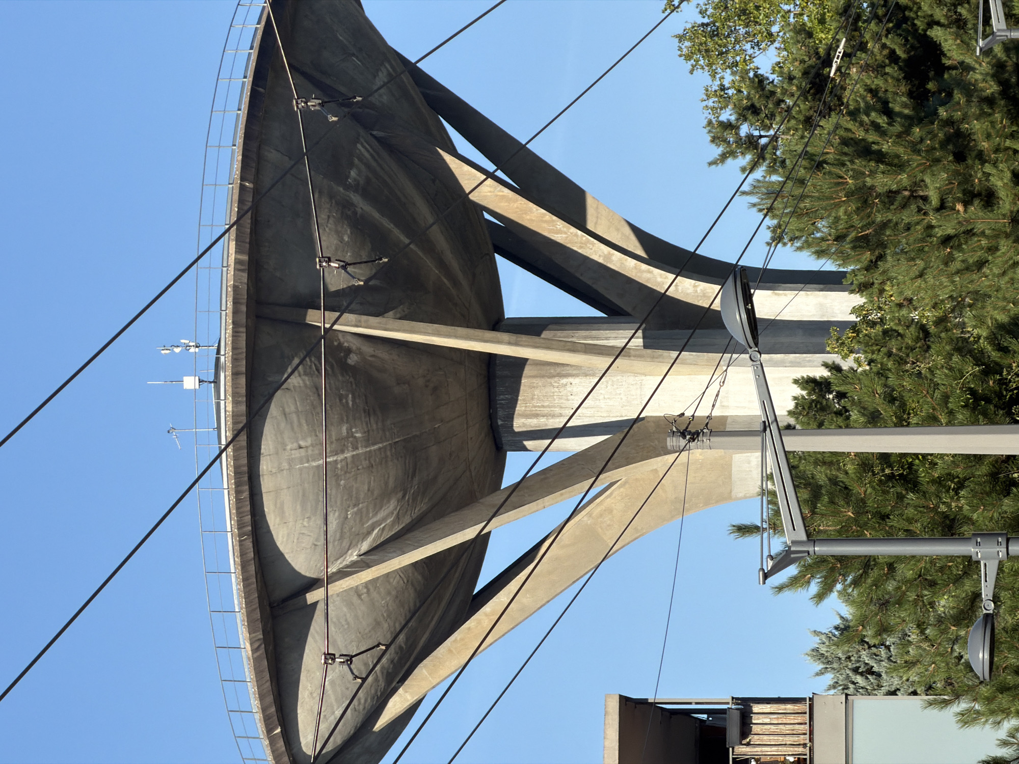 This is a full-shot, eye-level, outdoor, daytime, medium-wide photograph of the Geneva Water Tower, located in Lyon, France. The tower is constructed of concrete and features a large, bowl-shaped reservoir supported by three large, angled pillars and a central cylindrical pillar. A metal railing encircles the top of the reservoir. Various antennas and communication equipment are visible on the roof of the reservoir. In the foreground, several power lines crisscross the frame. A modern building with a balcony and a street lamp are visible to the left of the water tower. The background consists of a clear blue sky and lush green trees.