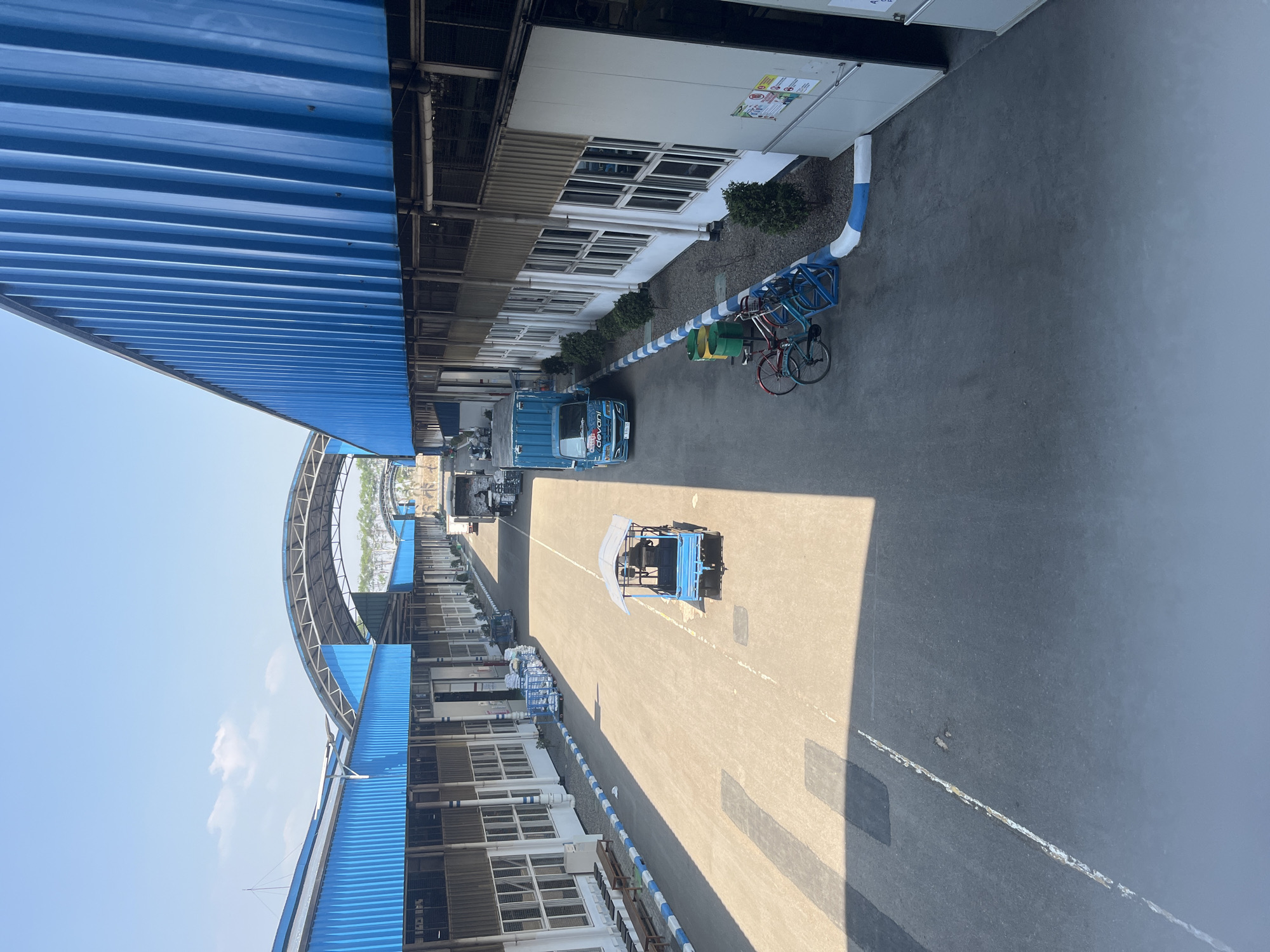 The image shows a view of a large industrial complex with blue corrugated metal roofs and white siding. The complex appears to be a warehouse or distribution center. In the foreground, there is a paved walkway with painted lines. A small blue electric cart with a canopy is parked on the walkway. Further down the walkway, there are several bicycles leaning against a blue and white striped pole. To the right, there is a blue truck with "Devani" written on the side. The complex is likely located in the city of Pelang, Indonesia, given the context. The environment appears to be an outdoor area with clear skies and sunlight casting shadows.