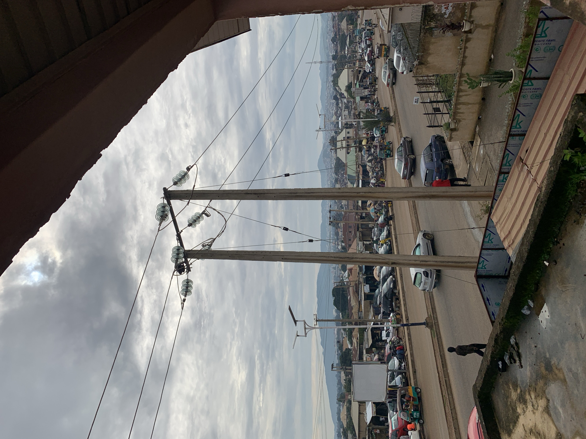 The image depicts an overcast sky over a bustling street scene in Rafin Par, Nigeria. A prominent power line with large ceramic insulators runs diagonally across the frame, supported by two concrete poles. The street is lined with various vehicles, including cars, motorcycles, and a three-wheeled auto-rickshaw, indicating active traffic. Buildings with tin roofs and simple facades flank the road, suggesting a commercial or residential area. A few pedestrians are visible, some walking along the sidewalk, others interacting with vendors or vehicles. In the background, a hilly landscape is visible under the cloudy sky, with more urban development stretching into the distance. The foreground shows a partially visible concrete structure, possibly a rooftop or balcony, with some debris scattered on its surface.