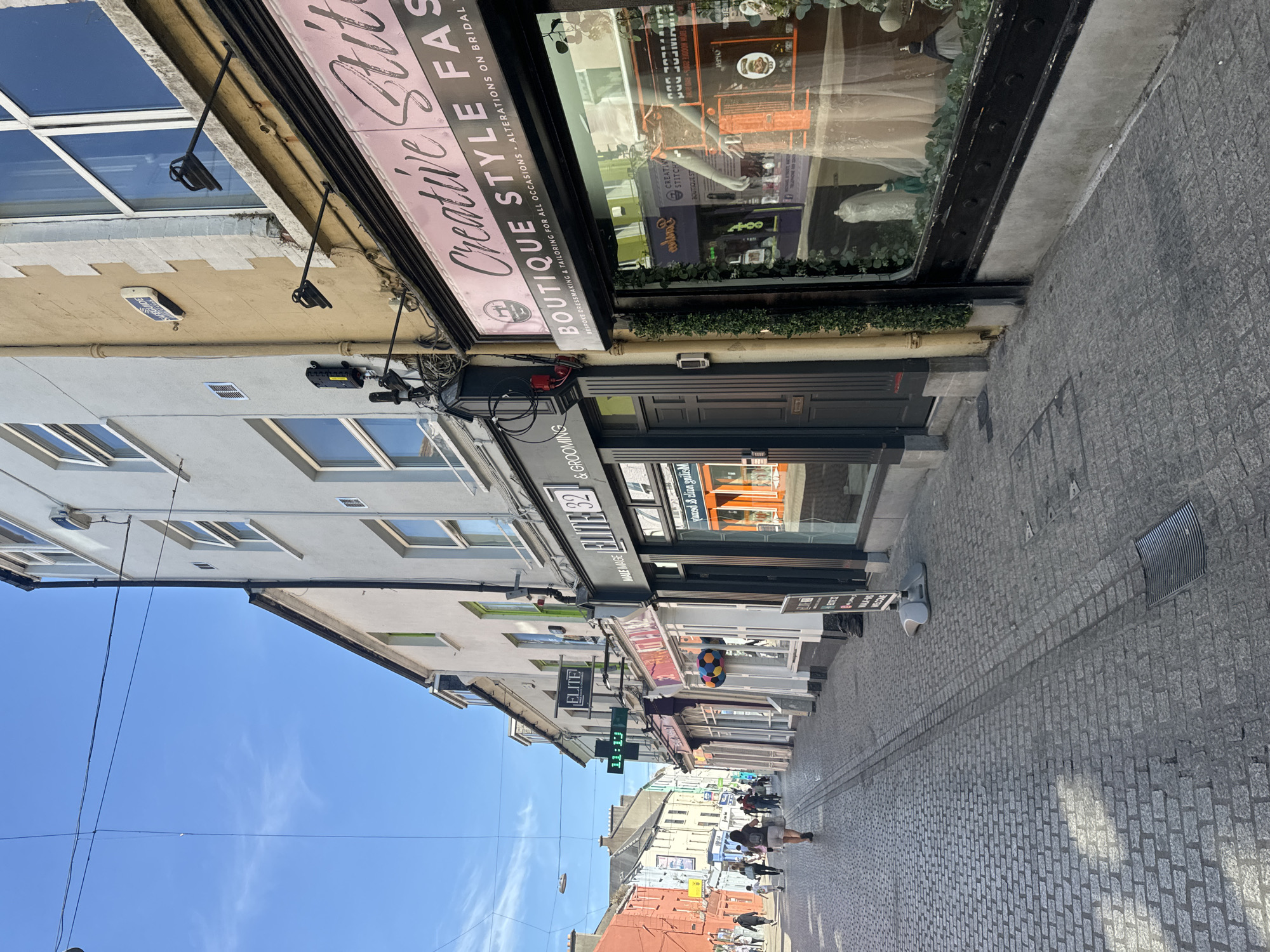 This is a street-level, outdoor, medium shot of Georges Street in Waterford, Ireland, taken on a sunny day with a clear blue sky. The cobblestone street is lined with shops and buildings. In the foreground, the street is paved with grey cobblestones, with some light and shadow patterns visible. To the right of the street is the facade of a building with a shopfront displaying a mannequin in a wedding dress in its window. The shop's sign reads "Creative Style Fashion Boutique", with smaller text beneath indicating "Bespoke Dressmaking & Tailoring for All Occasions - Alterations on Bridal". Next door, a shop labeled "Elite 32 & Grooming" is visible. Further down the street, the colorful facades of more buildings can be seen. Several people are walking along the street, including a woman in the foreground walking away from the camera. The scene captures a typical day in a bustling Irish city center.