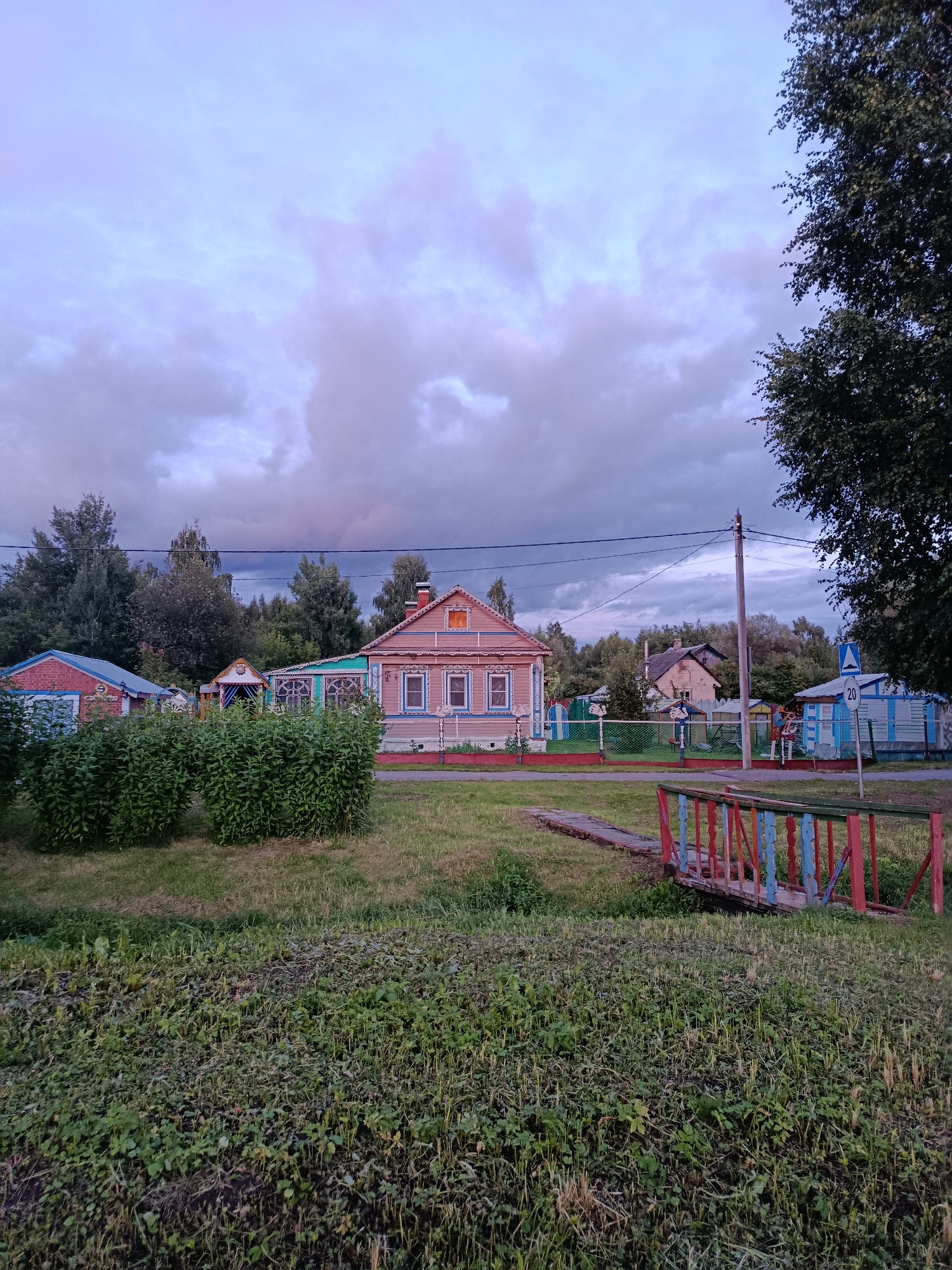 The image depicts a rural scene in the city of Veriya, Russia, under a cloudy sky with purple and gray hues. In the foreground, a patch of overgrown grass and weeds transitions into a mown lawn. A small, rustic bridge with red, blue, and green painted railings crosses a dry ditch or shallow waterway. Beyond the bridge, a row of dense green bushes lines a grassy area.
Further back, several houses are visible. The most prominent is a light pink wooden house with ornate white trim around the windows and a decorative gable. To its left is a smaller brick building with a blue roof. Behind these, a light blue wooden structure and another house with a gray roof are partially obscured by trees. A chain-link fence runs across the middle ground, separating the properties. A utility pole with overhead wires stands near the center of the image. A blue road sign with a white circle and "20" is visible to the right. The background is filled with a dense line of green trees. The overall impression is one of a quiet, perhaps early evening, rural setting.