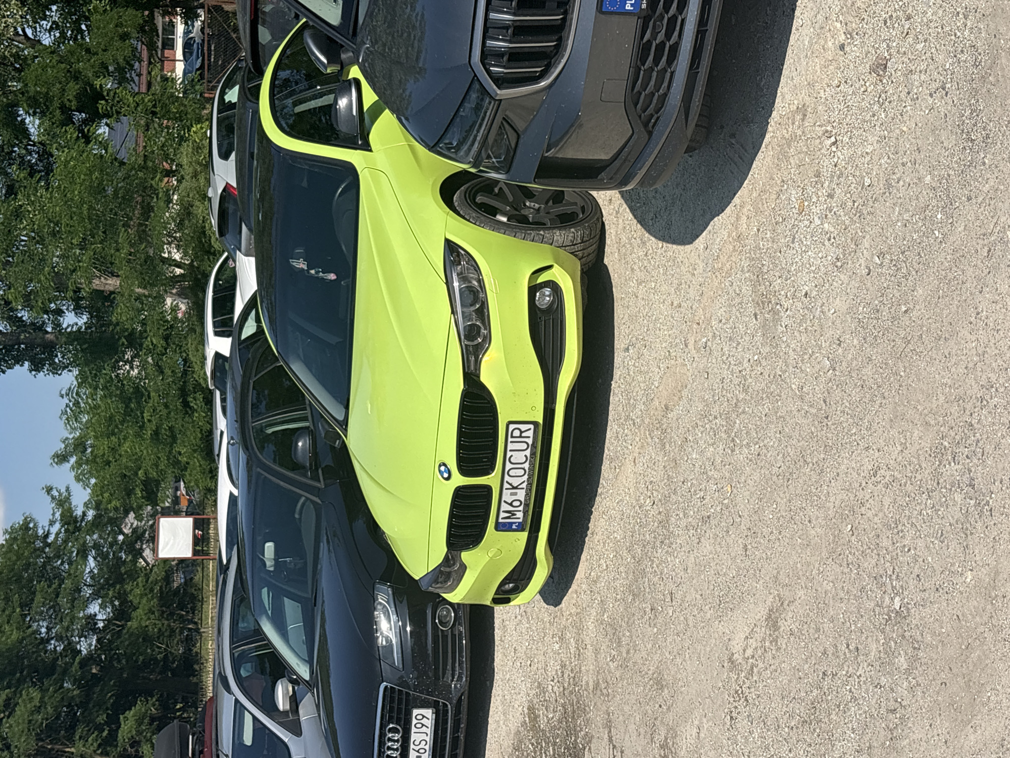 In Kobyla Góra, Poland, a bright lime green BMW coupe is parked on a gravel lot next to a black Audi sedan and a dark gray SUV. The BMW's license plate reads "M6-KOCUR" with the text "GRUPA SLIWINSKI" underneath. The Audi's license plate is partially visible, showing "6SJ99". In the background, trees and buildings are visible under a clear blue sky. The scene appears to be a daytime outdoor setting.