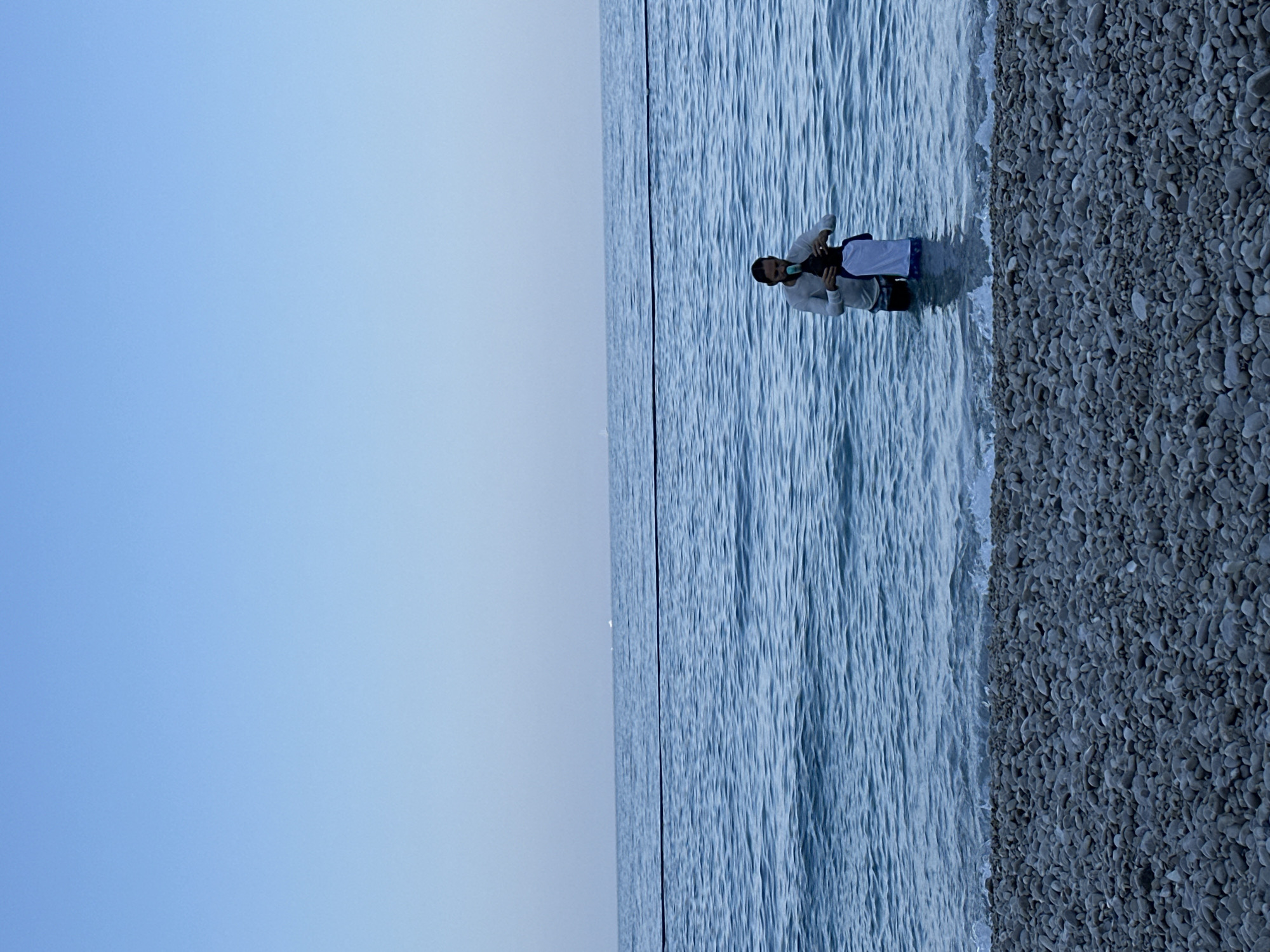 A man and a child stand in the shallow, blue water of the sea at a pebble beach. The man, wearing a long-sleeved shirt and shorts, is holding a blue object near the child's head. The child, wearing a white t-shirt and patterned shorts, is facing away from the viewer, towards the man. The water is calm with small waves breaking on the shore. The sky is clear and pale blue, with a faint horizon line visible in the distance. A dark line stretches across the water, likely a rope or buoy line. The pebble beach is made up of many small, smooth stones. The scene suggests a peaceful moment at a coastal location, possibly in Fethiye, Turkey, known for its pebble beaches and clear waters.