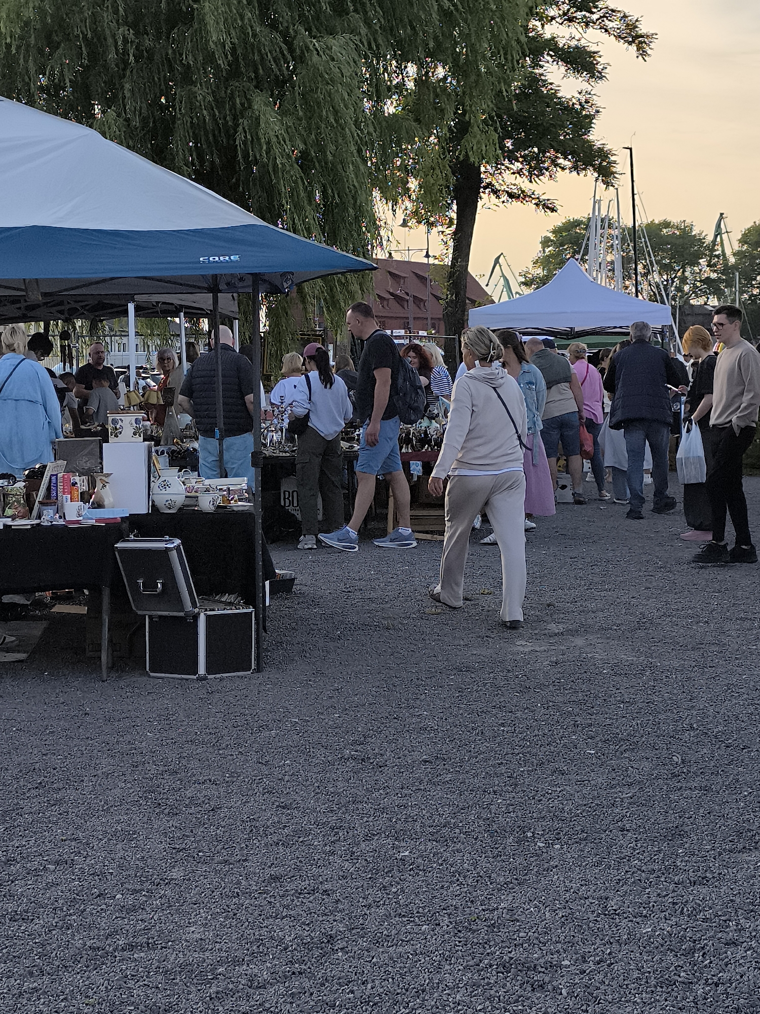 At dusk, a bustling outdoor market scene unfolds in Klaipėda, Lithuania, likely near the Old Town or a waterfront area given the presence of nautical elements in the background. The foreground is dominated by a vendor's stall laden with various items, including ceramics, books, and what appear to be decorative boxes. A large, dark-colored portable cooler with silver accents sits open on the ground beside the stall, suggesting ongoing sales or preparation.
A diverse crowd of individuals, seemingly shoppers and vendors, are gathered around the stall and milling about the market. Many are casually dressed, engaged in browsing or conversation. Among them, a woman in a light-colored tracksuit walks away from the camera, her back to the viewer. To her left, a man in a dark jacket and blue jeans stands observing the scene.
The market is set up under a blue and white striped tent canopy, providing shade. In the background, the warm glow of the setting sun illuminates the sky, casting a soft light on the scene and highlighting the silhouettes of trees and distant industrial structures, possibly cranes or parts of a shipyard, reinforcing the port city setting of Klaipėda. The ground is covered in dark gravel.