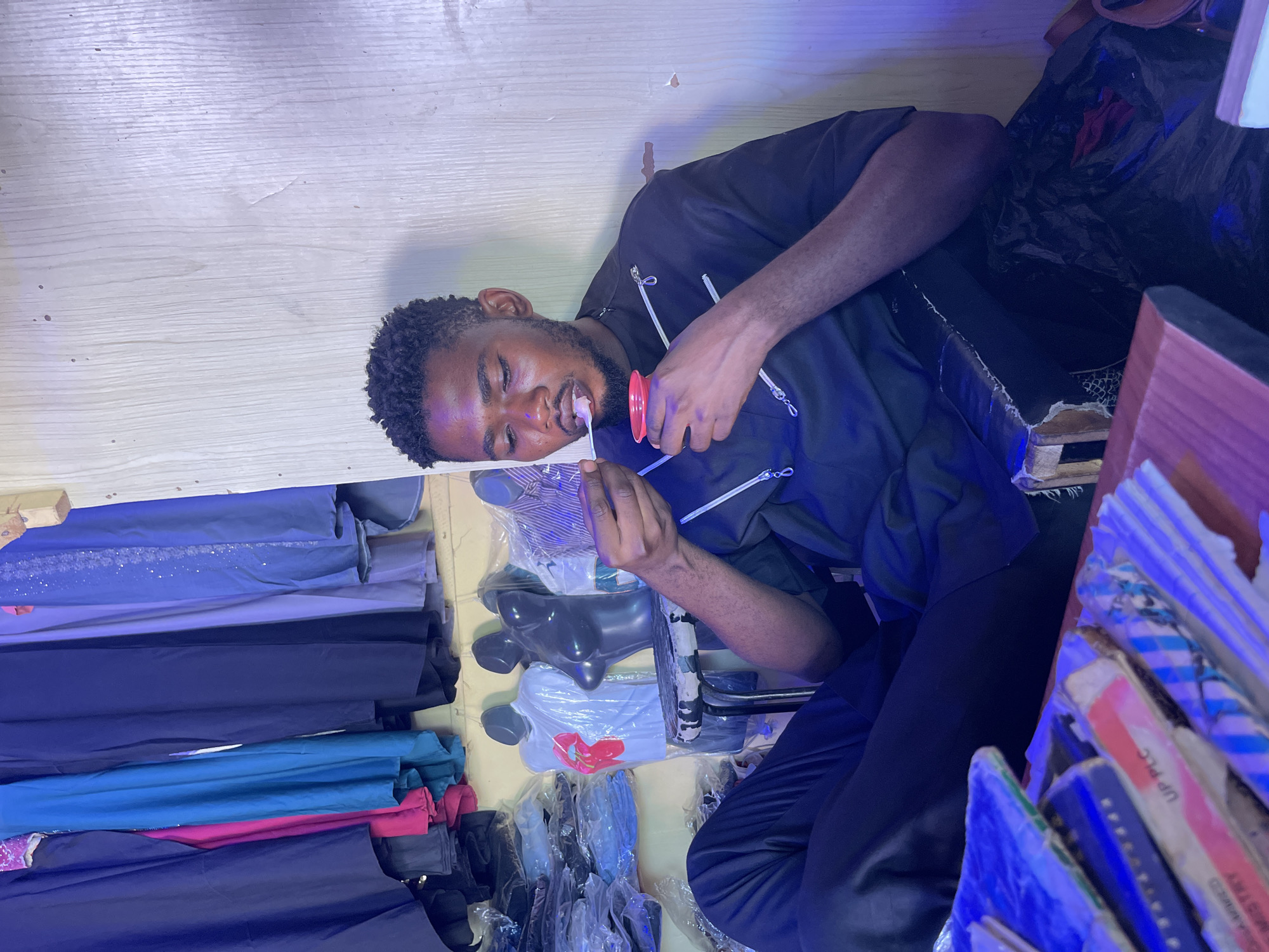An individual is seated amidst an array of textiles and apparel in Samaru, Nigeria. The person is consuming something from a red container using a utensil. Stacks of fabrics in various colors and textures are visible to the individual's left. Apparel on mannequins, some still in packaging, is also present. To the individual's right, there are stacked items, possibly books or packaging materials. The background suggests an indoor setting, perhaps a shop or storage area.