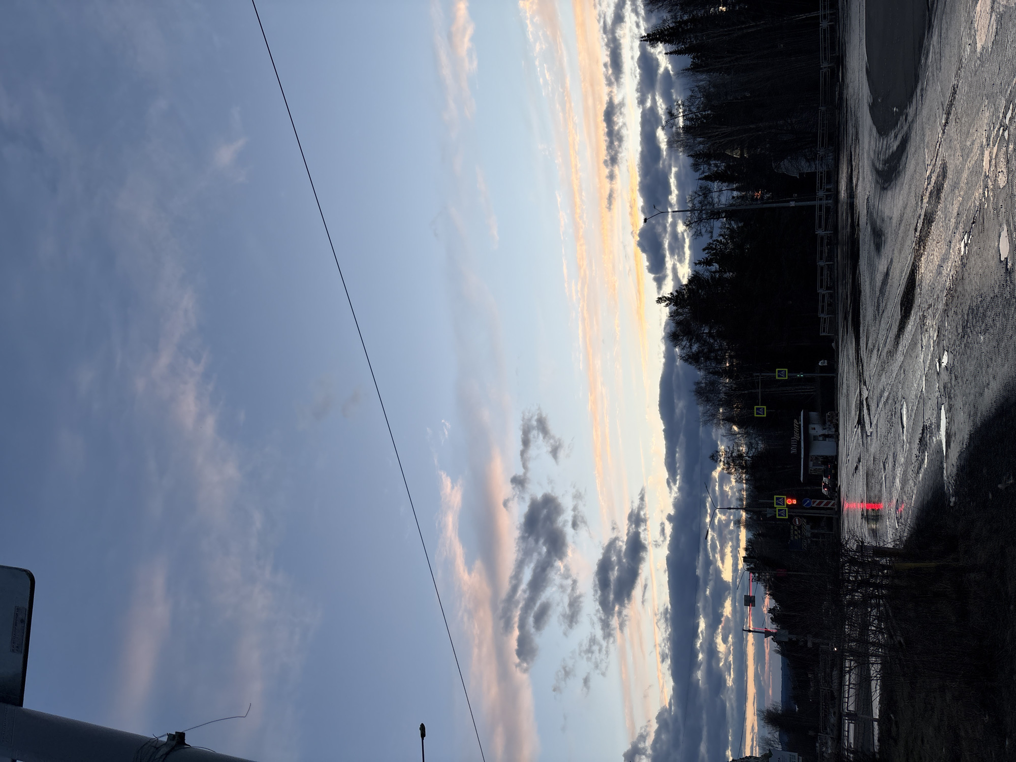 The image depicts an outdoor scene at dusk or sunset in Ust-Ilimsk, Russia, likely after recent rain given the wet, reflective road surface. The sky dominates the upper and left parts of the frame, showcasing a beautiful blend of light blue, soft pink, orange, and yellow hues, particularly where the setting sun illuminates the clouds along the horizon. Various cloud formations are present, from wispy cirrus-like clouds to more defined, layered cumulus clouds. A thin, dark cable or power line stretches diagonally across the sky.
On the right side, a dark, wet asphalt road or paved area with puddles reflects the sky's colors. The road appears worn, with visible cracks and rough patches. A dense line of silhouetted trees, possibly a forest, runs along the horizon, its dark branches stark against the colorful sky, suggesting late autumn or winter.
Amidst the trees and structures, several details are visible: a traffic light displays red signals, and a yellow diamond-shaped road sign indicates a pedestrian crossing. Behind the trees, a building or structure features a yellow sign with prominent Cyrillic text reading "ДОМ ТЕАТРА" (House of Theater). A street lamp or utility pole is also silhouetted against the sky. No people or specific activities are visible, contributing to a serene and somewhat muted atmosphere as day transitions to night.