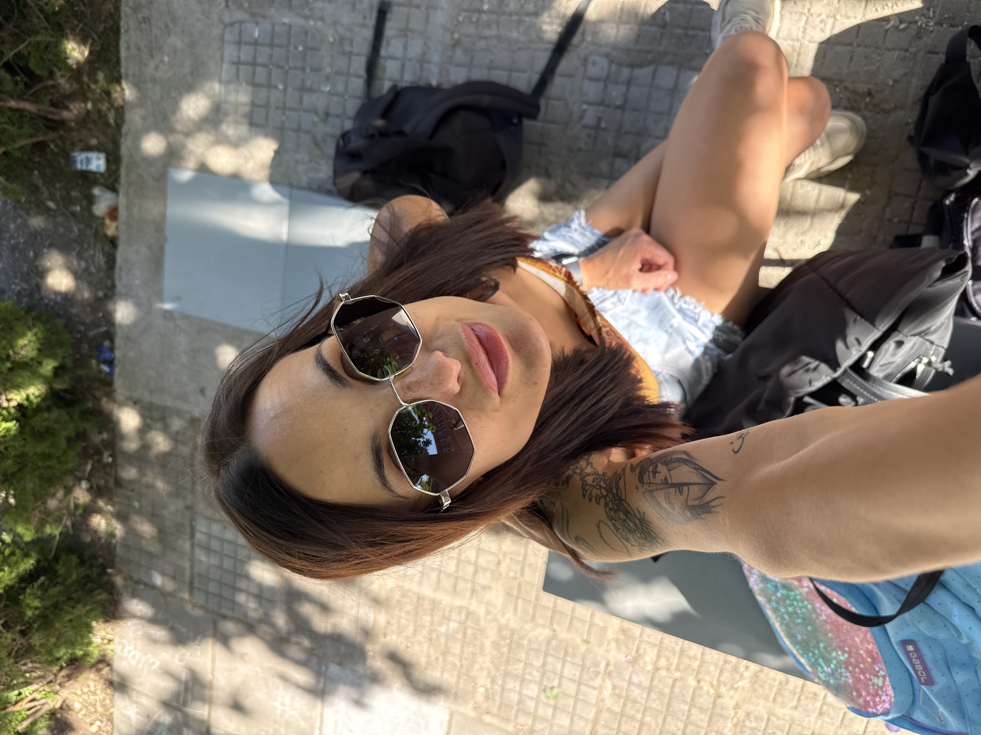 A selfie-style, top-down shot shows a woman in Pleven, Bulgaria. She's wearing sunglasses and a light-colored top, seated with her legs crossed and adorned with tan sneakers, surrounded by bags and a small bush. The location appears to be a paved area in Pleven, with light and shadow patterns suggesting a sunny day. Tattoos are visible on her arm.
