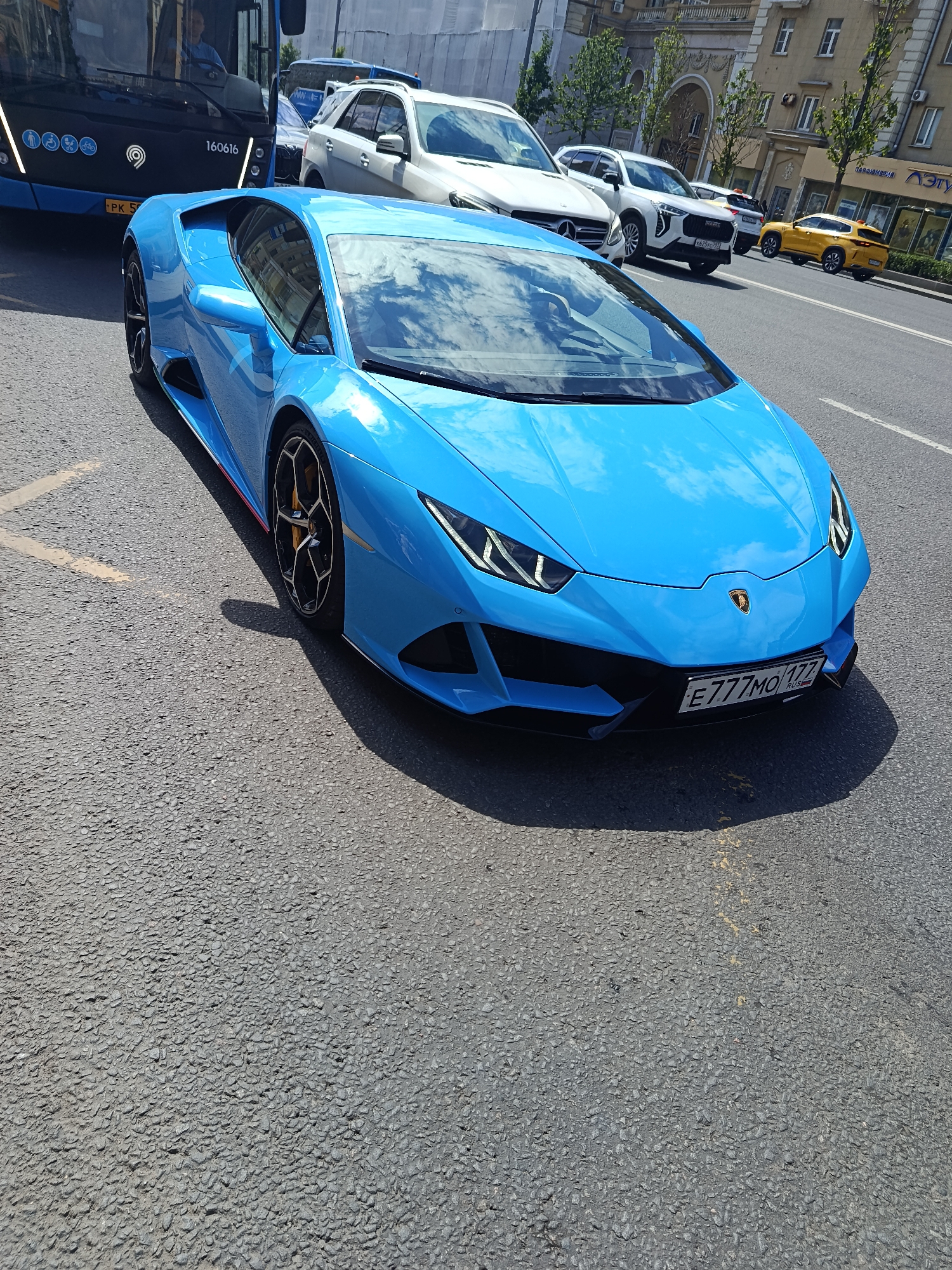A vibrant, bright blue Lamborghini Huracan is the central focus, positioned on a coarse asphalt city street. The luxury sports car is angled slightly, with its front facing towards the right side of the frame. Its glossy surface reflects a clear, sunny sky with scattered white clouds. The car features intricate black wheels with yellow brake calipers visible behind them. A Russian license plate, "E777MO 177 RUS", is affixed to its front bumper.
The scene depicts a busy urban environment, suggesting the car is either in traffic or briefly stopped. To the left and behind the Lamborghini, a dark blue city bus is partially visible. The bus displays the number "160616" and several accessibility icons, with its driver visible through the windshield in a light blue shirt. A line of other vehicles extends down the street to the right, including a white Mercedes-Benz SUV, a white Haval SUV (with license plate "X625PC 799"), and a distinct bright yellow SUV further in the distance.
On the right side of the street, multi-story buildings with classic architectural details and trees line the sidewalk, indicating a historic urban setting. A shop sign with partially visible Cyrillic text, "АЭТУ", is discernible on one of these buildings. The strong shadows cast by the vehicles and buildings suggest it is daytime, likely mid-morning or afternoon, under clear weather conditions. The presence of Russian license plates, Cyrillic text, and the architectural style confirm the location as Moscow, Russia.