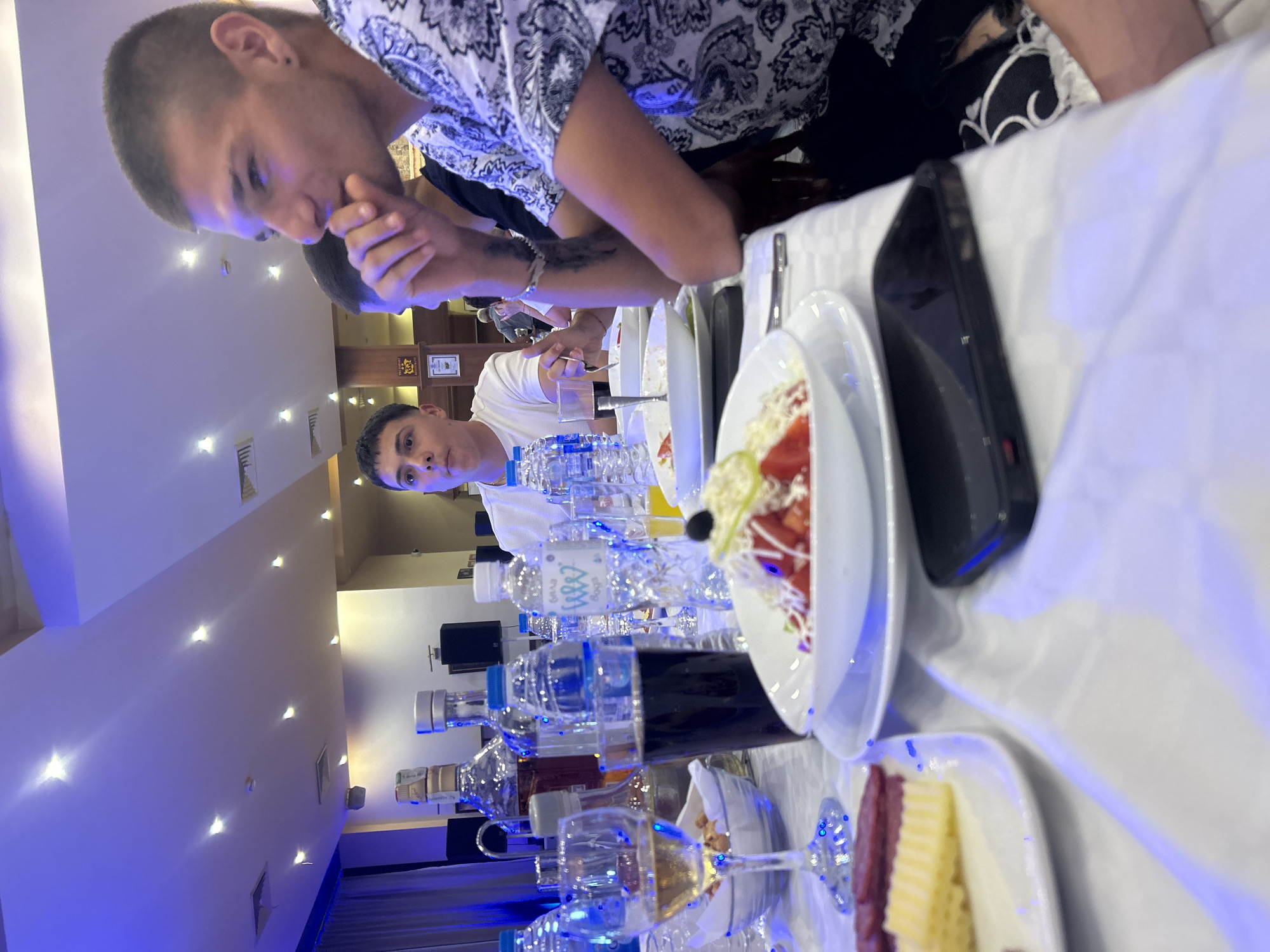 The image shows a long table laden with bottles of water, glasses, plates of food (salad and what appears to be cheese and meat), and a cell phone. Several people are seated at the table, seemingly eating or preparing to eat. The lighting suggests an indoor setting, possibly a restaurant. The presence of water bottles and the types of food indicate a meal or a gathering with refreshments. The picture was taken in a restaurant of the city Elhovo, Bulgaria.