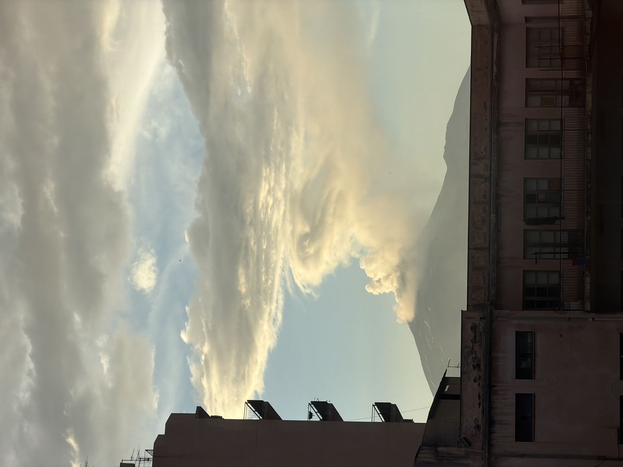 The image depicts a view of Mount Etna erupting, viewed from the city of Catania, Italy. In the foreground, the upper floors of two buildings are visible. The building on the left is tall and has multiple balconies with metal railings. The building on the right is lower and appears older, with a pink facade and many windows. The erupting volcano is in the background, with a large plume of smoke and ash rising into the sky. The sky is filled with clouds, some of which are illuminated by the sun. The overall impression is one of a dramatic natural event occurring within an urban setting.