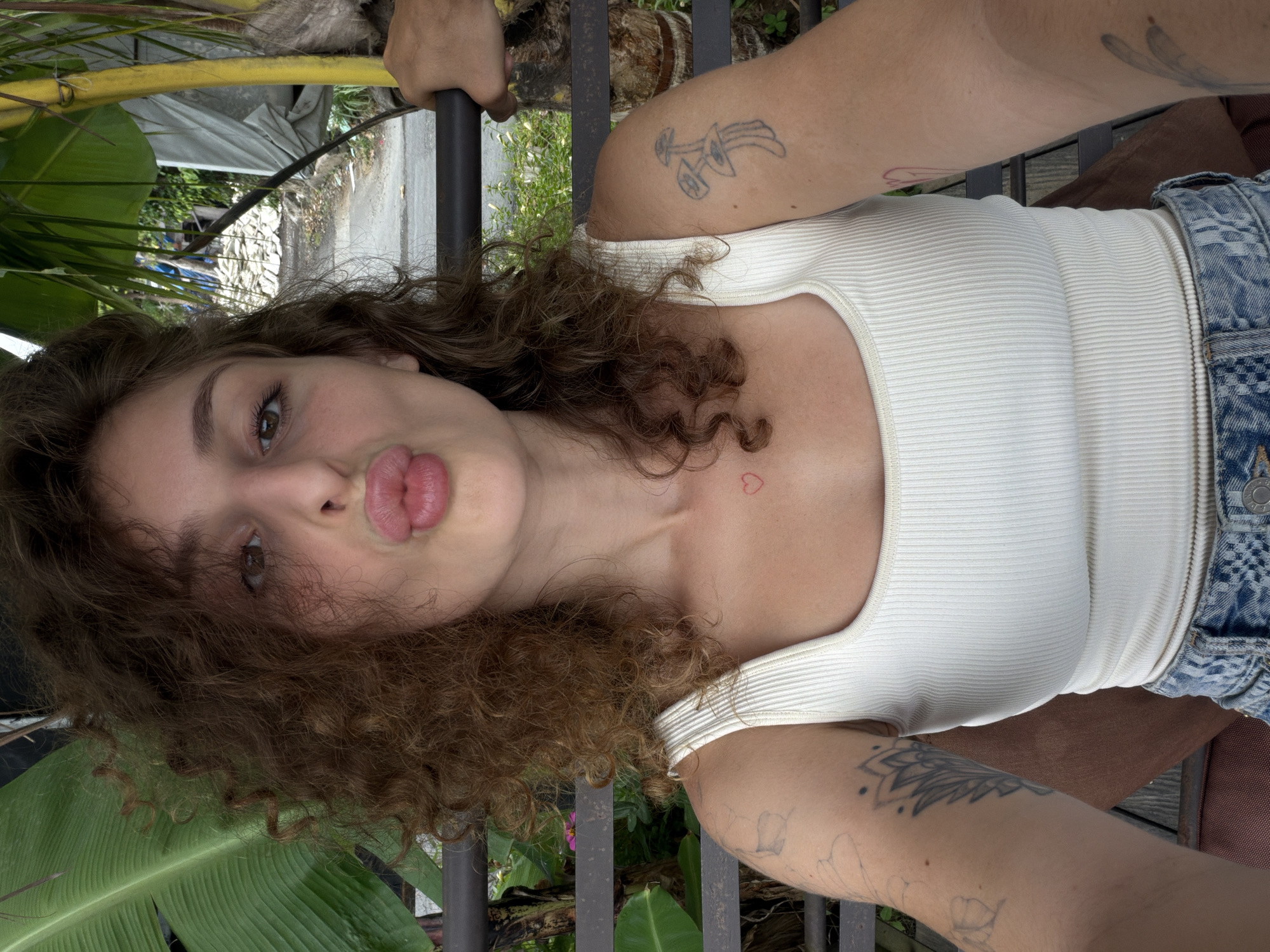A young woman with curly brown hair and light brown eyes is taking a selfie. She is wearing a white ribbed tank top and denim shorts. Her lips are pursed, and she has a small red heart tattoo on her chest. She has tattoos on her arms, including a mushroom tattoo on her right arm and a floral tattoo on her left arm. She is sitting on a metal bench outdoors, with lush green foliage and a dirt path visible in the background. The scene appears to be in a tropical or subtropical environment, possibly a garden or a pathway in a village. Given the context provided, the location is in the city of Mengwi, Indonesia.