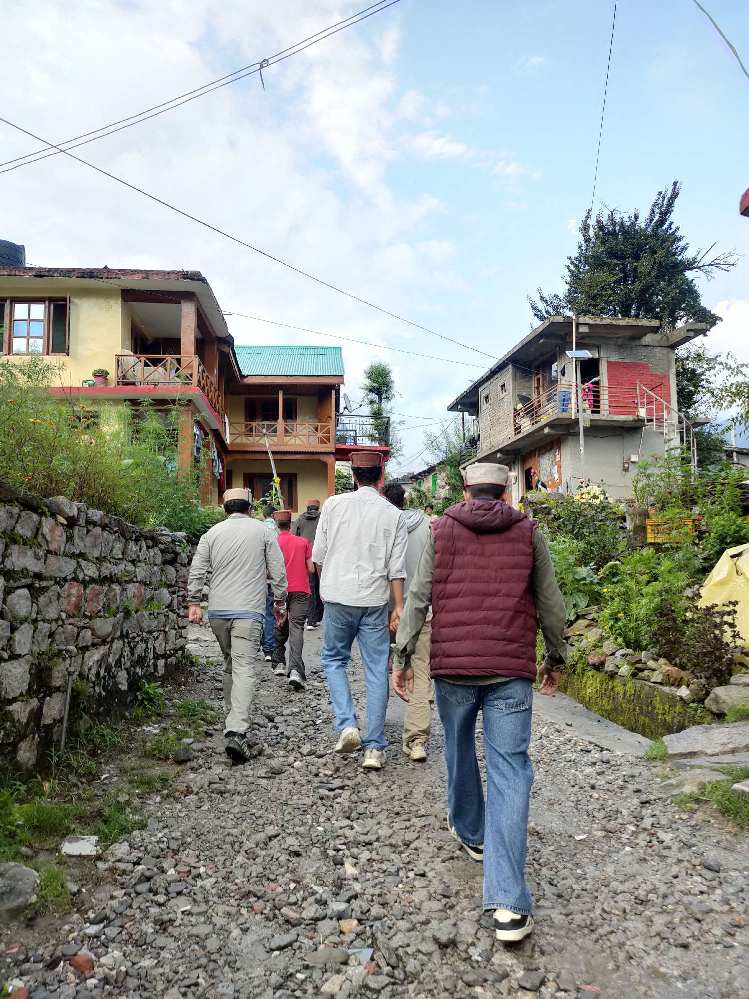 A group of men are walking away from the camera down a narrow, rocky street in Burwa, India. The men are dressed in casual clothing, with most wearing traditional caps. The street is lined with stone walls and houses, some of which are brightly colored. Lush green vegetation grows along the sides of the street and between the houses. The sky is cloudy, with patches of blue visible. The scene appears to be a rural or semi-urban setting.