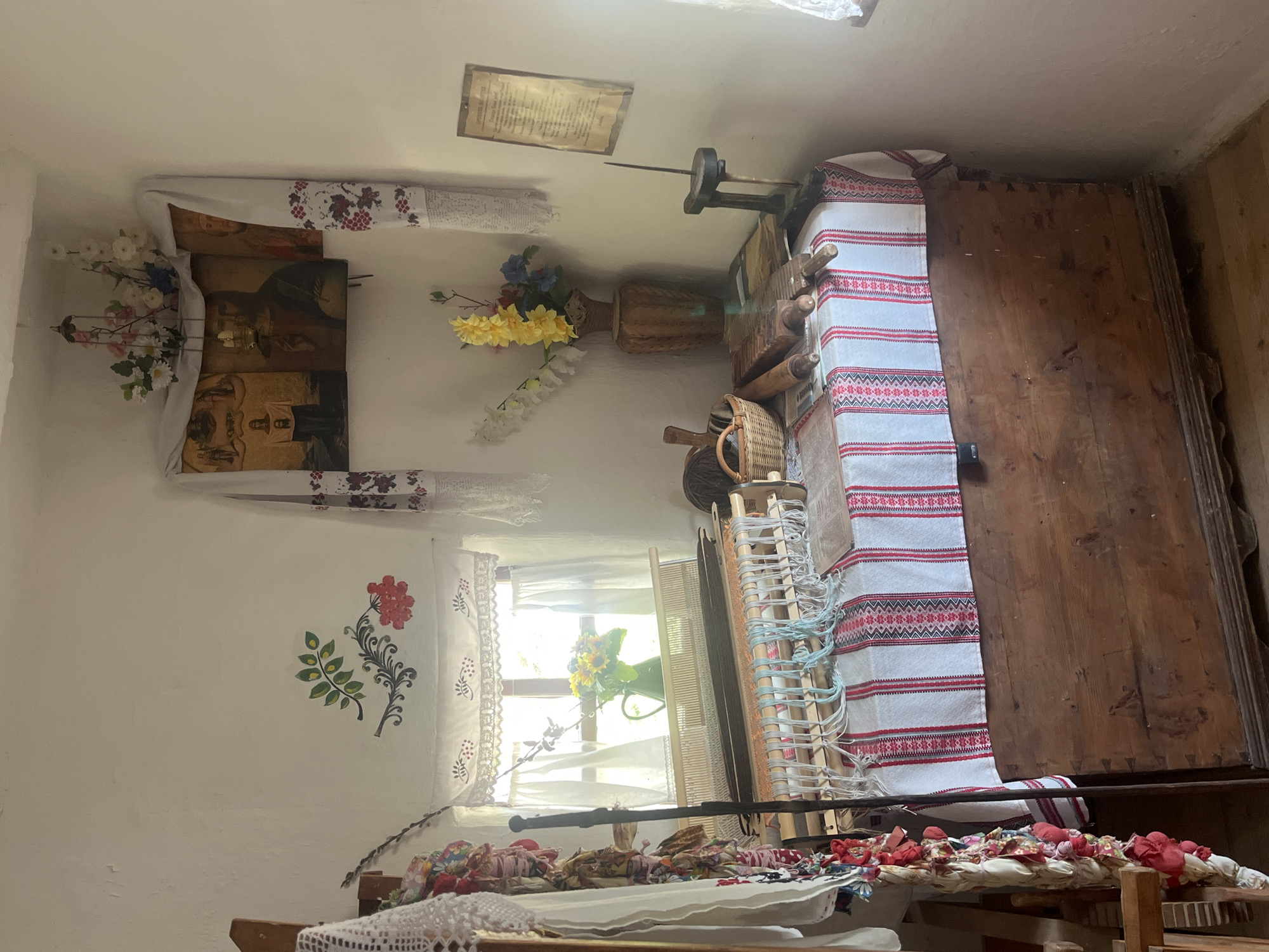 This image depicts an interior scene inside what appears to be a traditional dwelling or museum exhibit in Taman', Russia. The room has white, textured walls adorned with folk art motifs, including a branch with red berries and green leaves on the left wall, and a decorative embroidered cloth hanging over a religious icon in the corner. Sunlight streams through a window covered by white lace curtains, illuminating a pitcher of sunflowers on a loom.
The focal point of the room is a large wooden chest, on top of which is a loom with colorful threads. Beside the loom, on the chest's surface, are various items related to textile work and possibly food preparation: a small wicker basket, wooden rolling pins, and other tools. To the right, on a table, a vase holds artificial flowers, and a framed document is mounted on the wall. The overall impression is one of a preserved, culturally rich domestic space, likely showcasing historical crafts and traditions of the region.