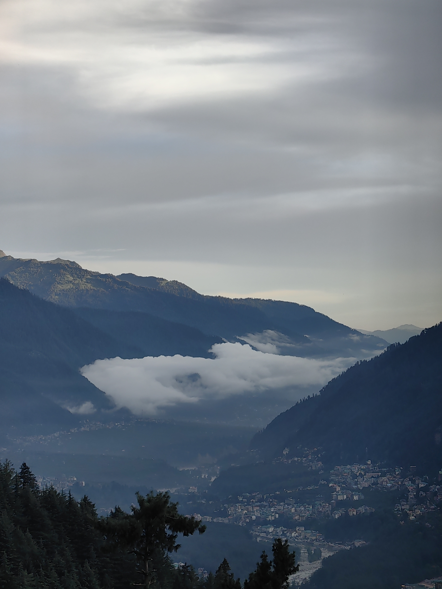 An elevated, wide-angle shot captures a misty mountain valley on a cloudy day. In the foreground, dark green pine trees line a steep slope, partially obscuring the view. Below, a town is nestled within the valley, its buildings a mix of white, grey, and muted colors, clustered along a river. Wispy white clouds drift through the valley, creating a hazy, dreamlike atmosphere. In the distance, layered mountain ranges rise, their peaks shrouded in mist and soft grey clouds. The overall impression is one of serene, natural beauty, with the town a small human presence within a grand, enveloping landscape.