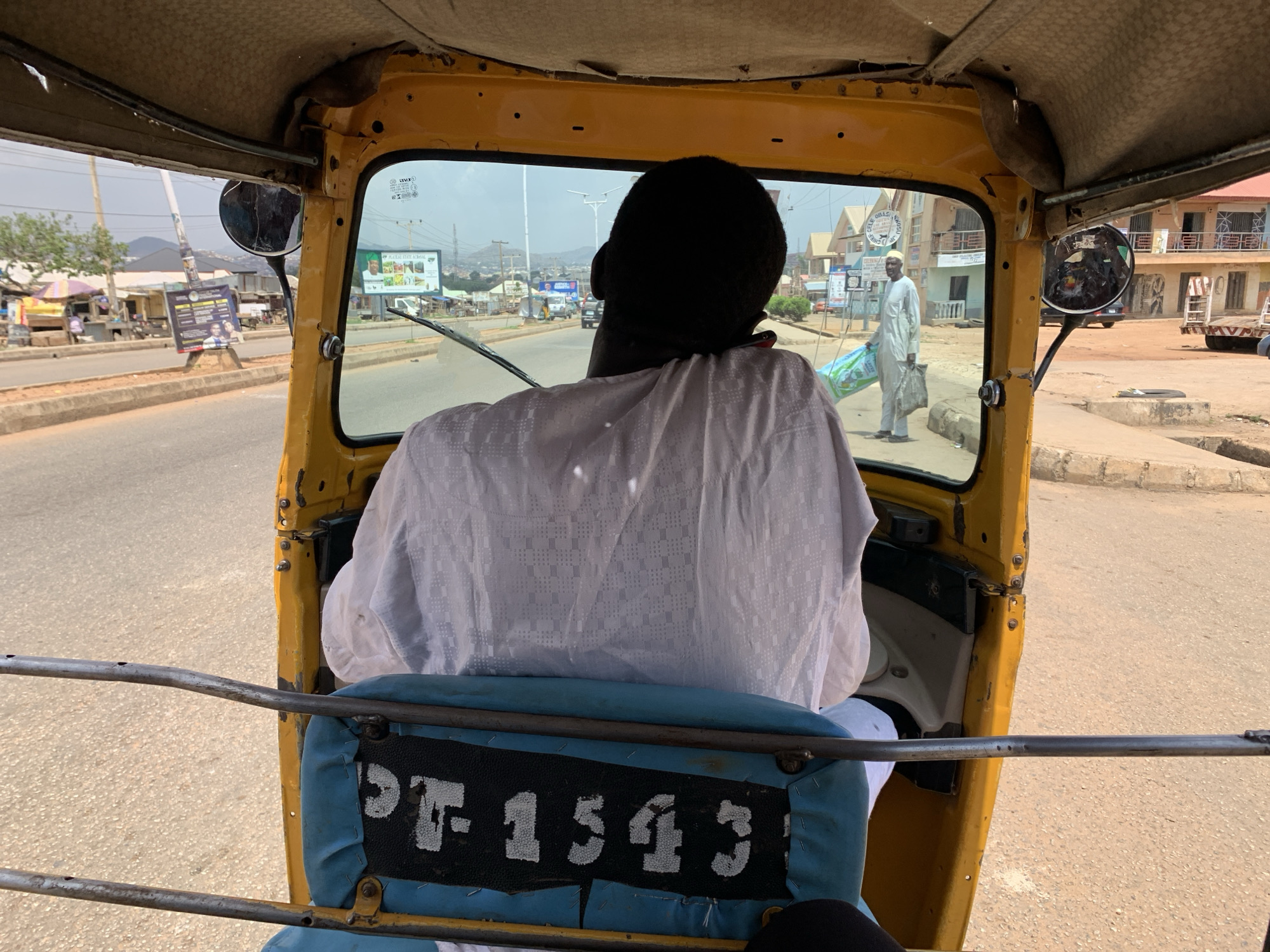 The image shows the interior of a yellow tuk-tuk, a three-wheeled motorized vehicle, with a view through the windshield onto a street scene in Rafin Par, Nigeria. The tuk-tuk driver, a Black man wearing a white shirt, is visible from the back. The blue seat behind the driver has "PF-1543" printed on it in white lettering. Through the windshield, a man dressed in white is visible on the right side of the street carrying a bag. Buildings are seen on both sides of the street, and a billboard advertising a "Plateau State Hospital" is visible in the distance. The road is paved and appears to be moderately busy.