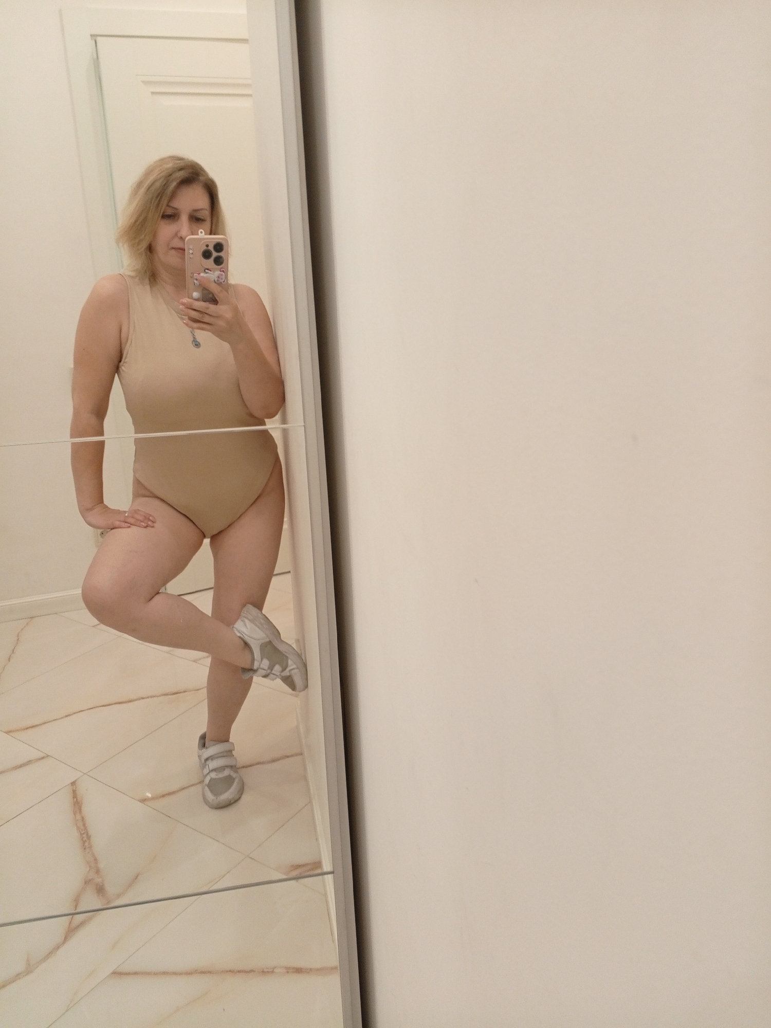 A woman is taking a mirror selfie in what appears to be a bathroom or dressing room. She is wearing a flesh-colored, one-piece garment that resembles a leotard or swimsuit, and grey sneakers with white soles and velcro straps. Her hair is blonde and shoulder-length. She is leaning against a mirrored wall, with her right leg bent at the knee and resting on her left leg. Her left hand is resting on her left thigh, and her right hand is holding a smartphone, which is capturing the photo. The mirror reflects her entire body and the tiled floor, which has a light beige color with marbled brown veins. To the right of the mirror is a plain, off-white wall. The overall lighting is bright and appears to be artificial, suggesting an indoor setting. There is no visible text in the image. The woman's expression is neutral. The setting is an interior, likely domestic, in Sochi, Russia.