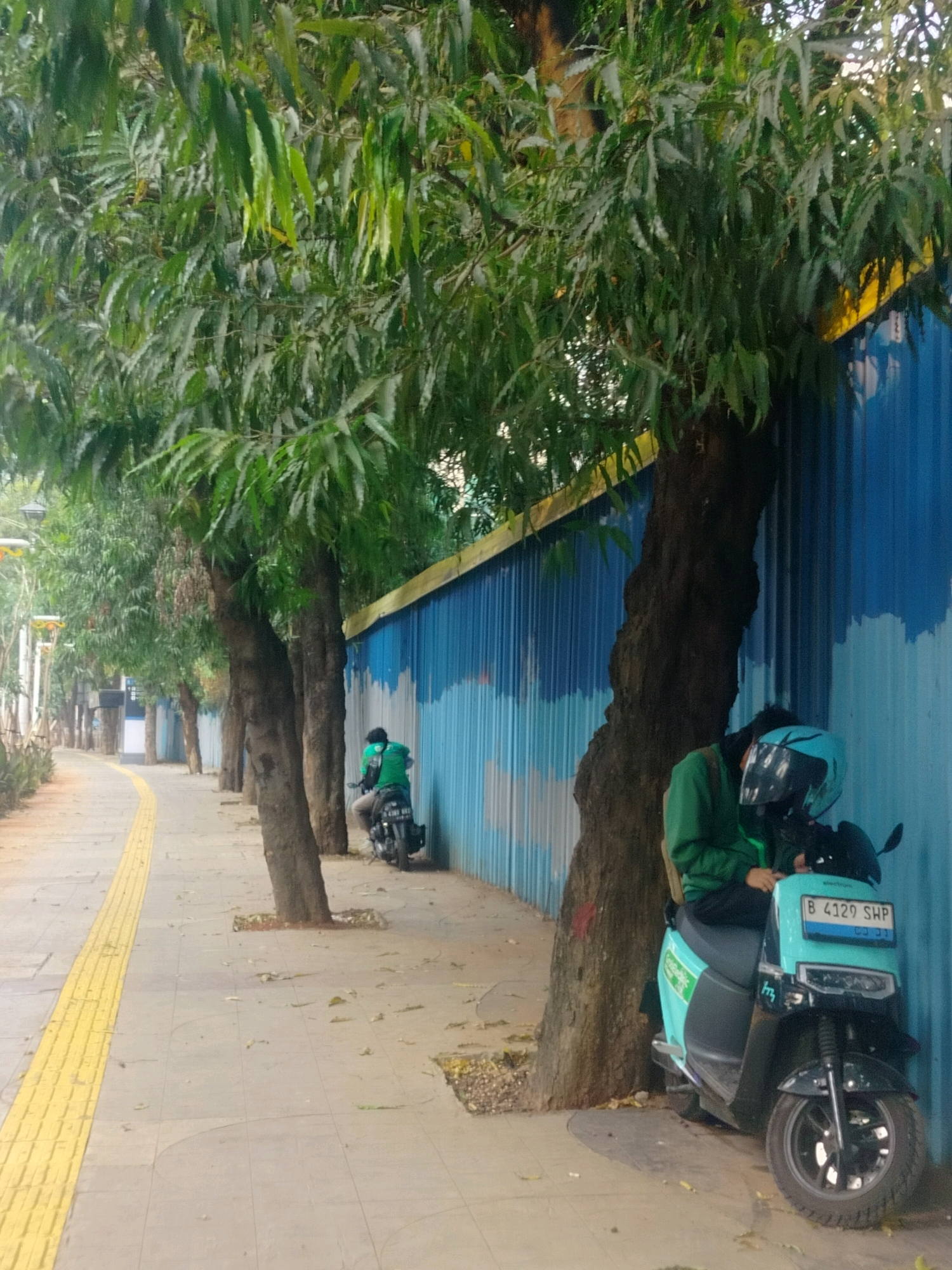 In the city of South Jakarta, Indonesia, a paved sidewalk with a tactile yellow warning surface runs alongside a row of trees. Behind the trees is a corrugated metal wall painted with blue and white waves. A person wearing a green jacket and helmet is seated on a turquoise electric scooter parked next to a tree. The scooter has a license plate that reads "B 4129 SHP". Further down the sidewalk, another person in a green jacket is riding a black motorcycle. Fallen leaves are scattered on the ground.