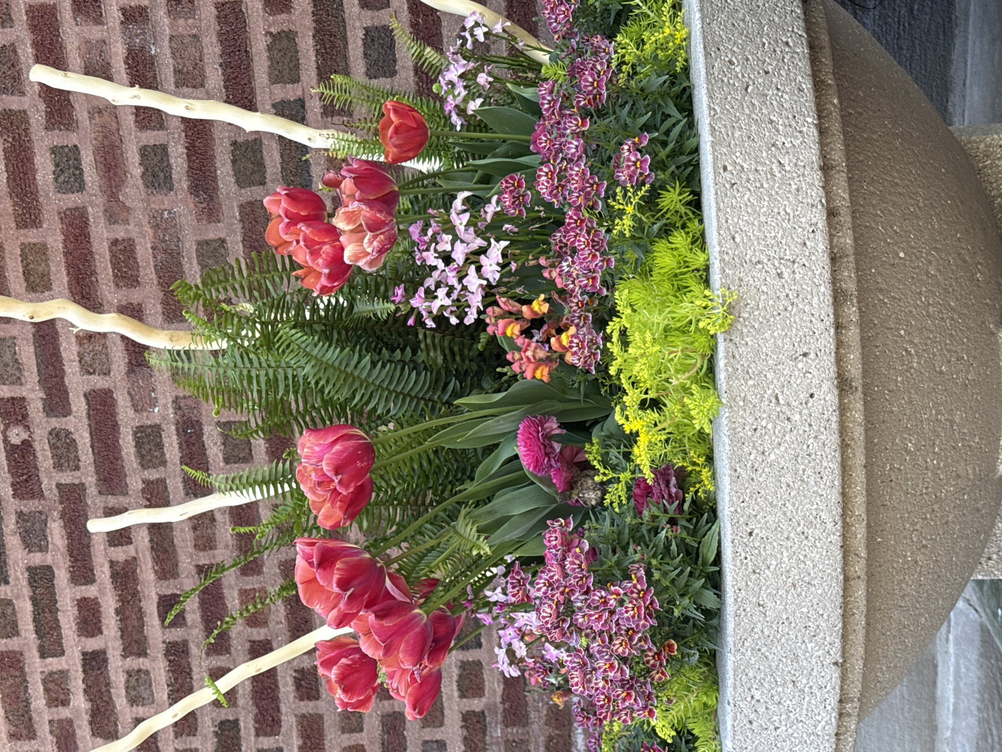 A vertical, eye-level shot shows a concrete planter filled with various flowers and greenery. The blooms include red tulips, purple and white orchids, and chartreuse-colored foliage. Three light-colored branches protrude from the planter. The planter sits against a brick wall. The location is likely Chicago, United States of America.